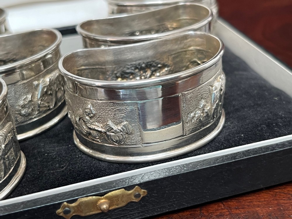 A cased set of six silver napkin rings with embossed relief scenes inside a black silk and velvet box. View VI