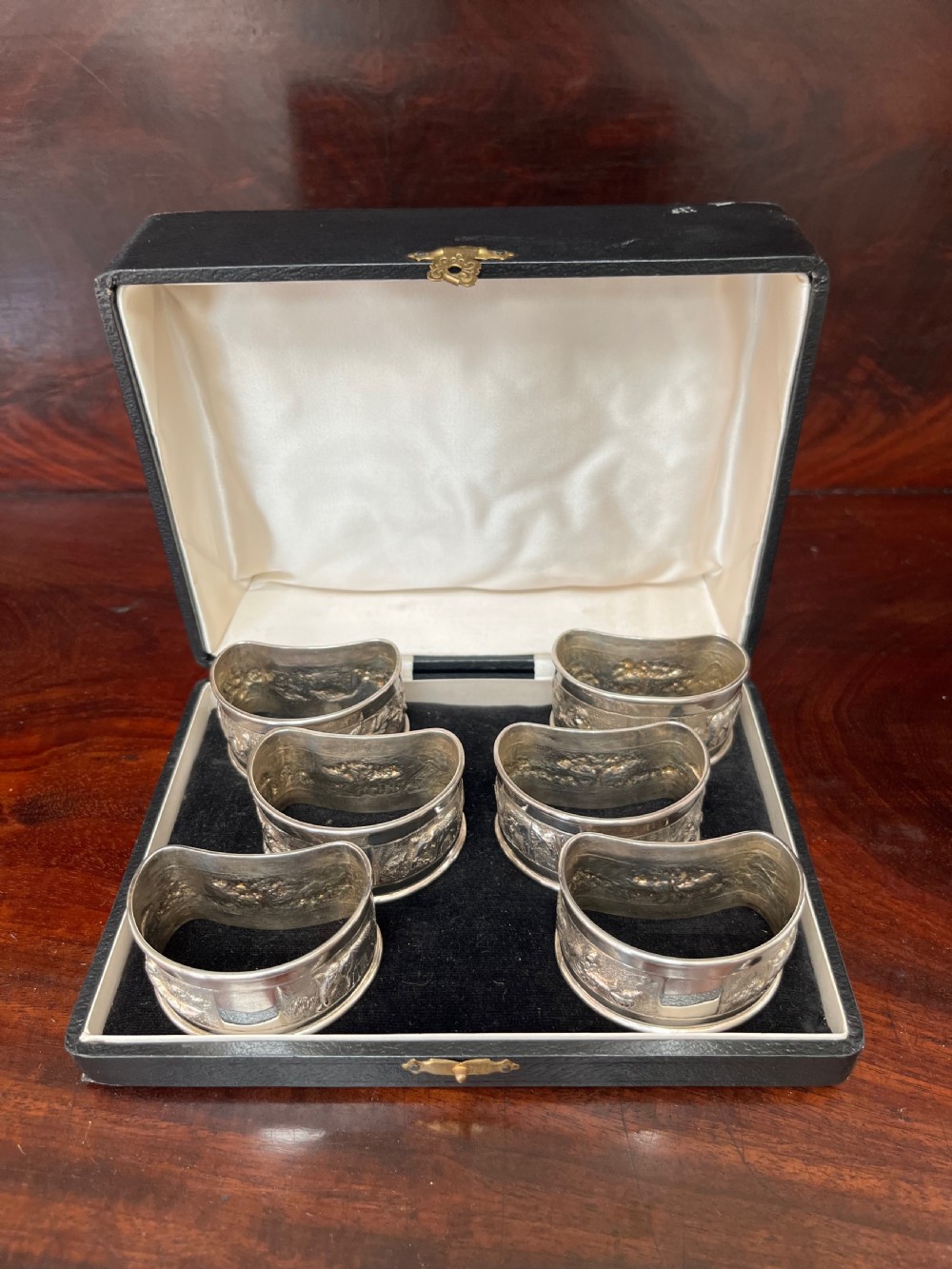 A cased set of six silver napkin rings with embossed relief scenes inside a black silk and velvet box. View VII
