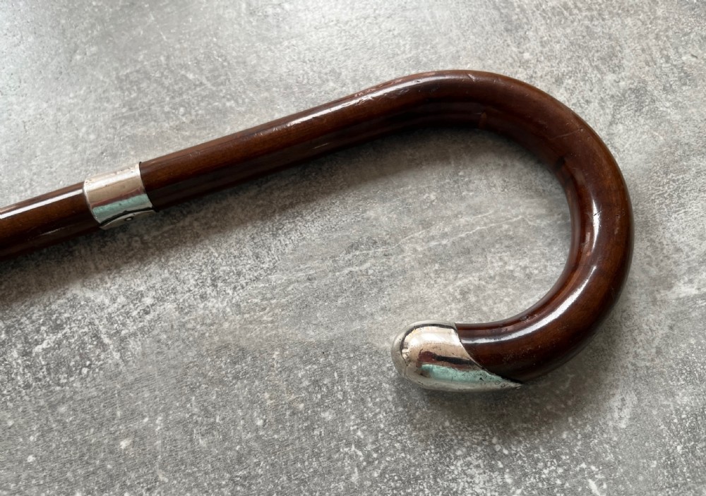A dark brown polished hardwood walking stick featuring a curved crook handle with silver mounts and a metal ferrule at the tip. View II