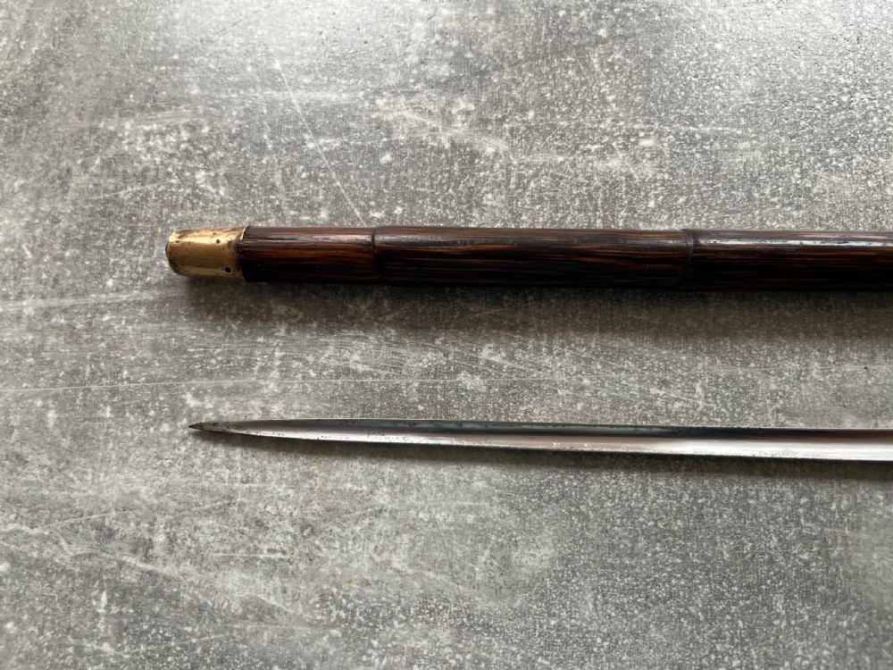 A dark wood walking stick featuring a crook handle and faux bamboo carved nodes, unsheathed to reveal a long steel blade alongside its wooden casing. View IX