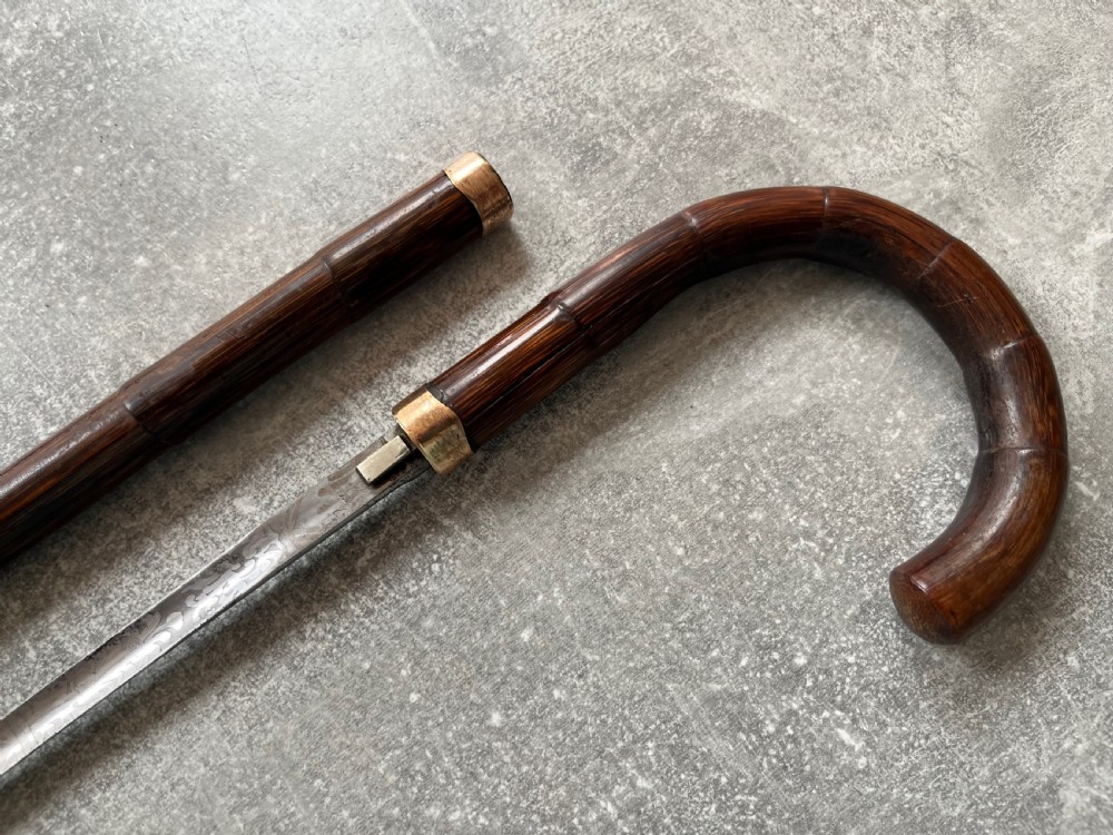 A dark wood walking stick featuring a crook handle and faux bamboo carved nodes, unsheathed to reveal a long steel blade alongside its wooden casing. View X