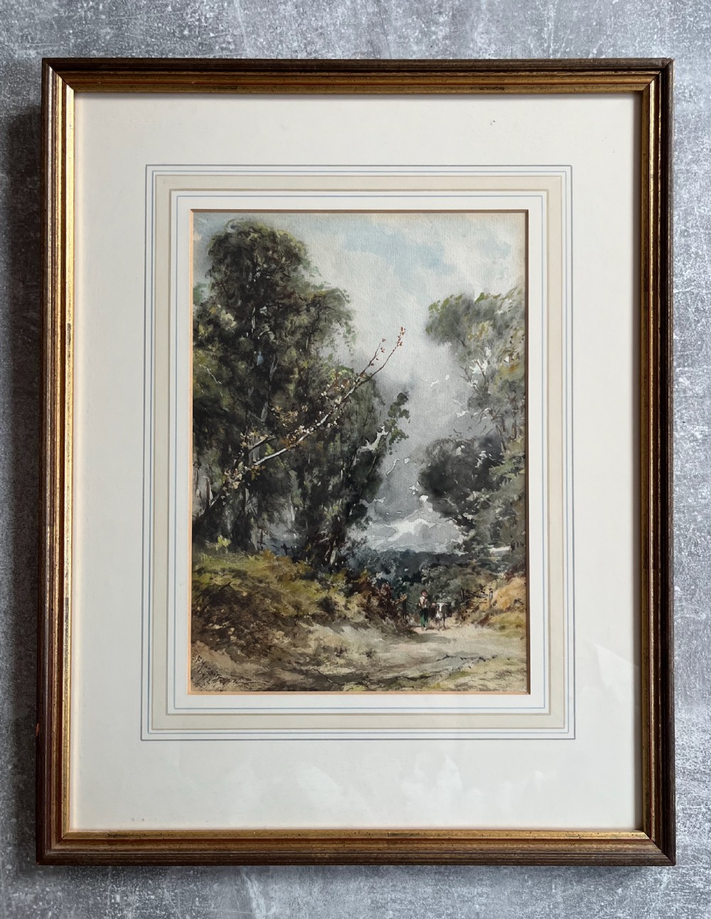 A framed 19th-century watercolour painting depicting a wooded countryside path under a clouded sky, mounted in a lined cream mat and narrow gilt-trimmed wooden frame.