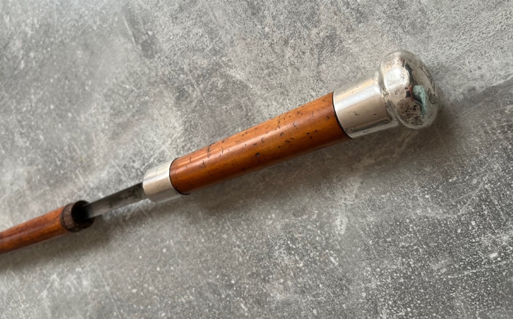 An antique malacca cane sword stick featuring a hallmarked silver pommel collar and an etched steel blade alongside its wooden sheath. View IV