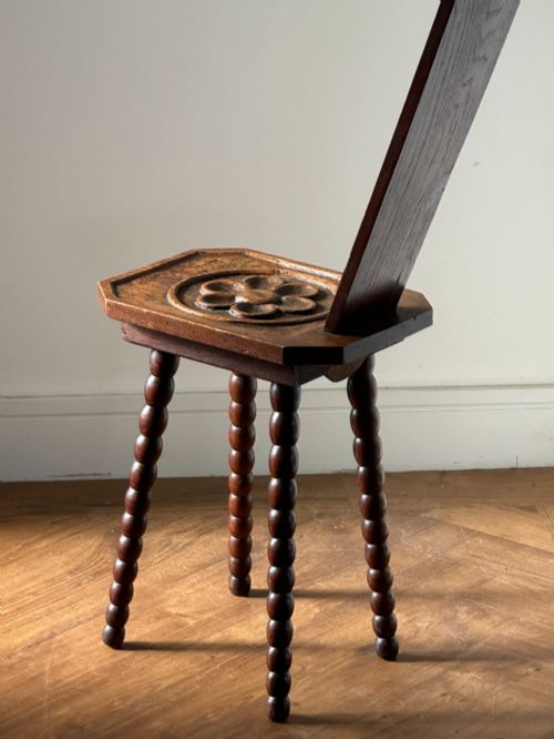 fig 1. An Arts and Crafts carved wooden spinning stool featuring a tall narrow backrest, a relief-carved floral seat, and bobbin-turned legs.