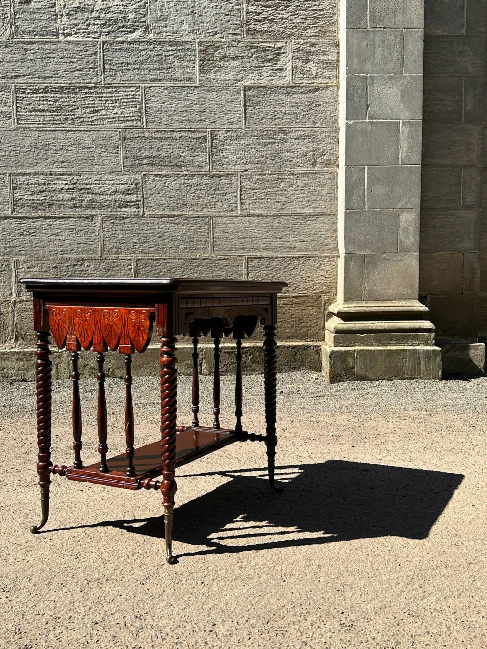 An Aesthetic Movement stained birch centre table featuring barley twist legs, carved foliage aprons, a lower tier with slender spindles, and brass sabots.