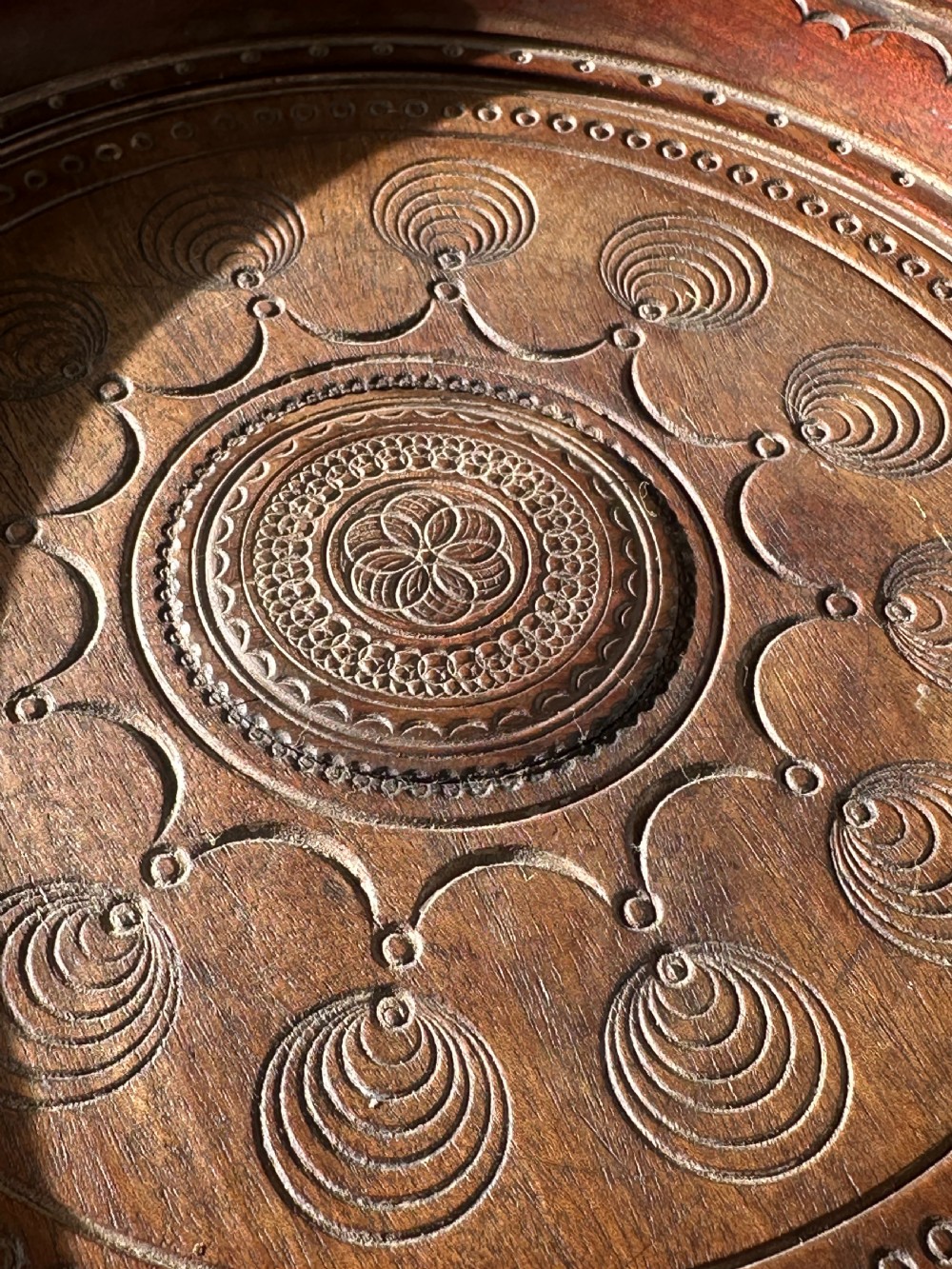 A circular carved mahogany decanter coaster featuring intricate central geometric patterns, a gadrooned rim, and small castors mounted underneath. View II