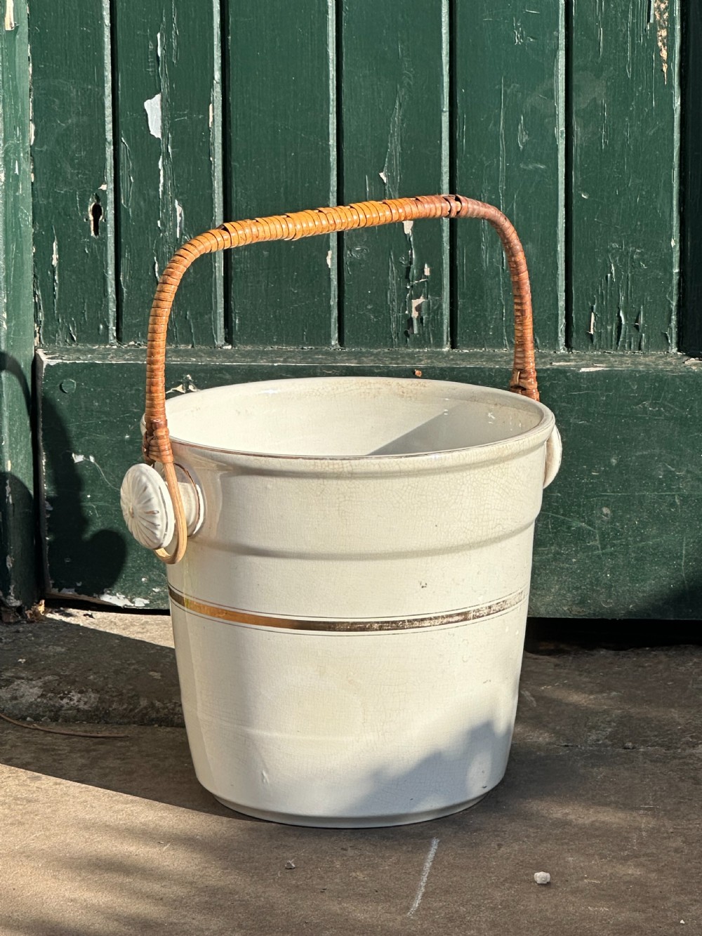 A cream-glazed ceramic bucket featuring a horizontal gilt line and a cane-wrapped overhead swing handle.