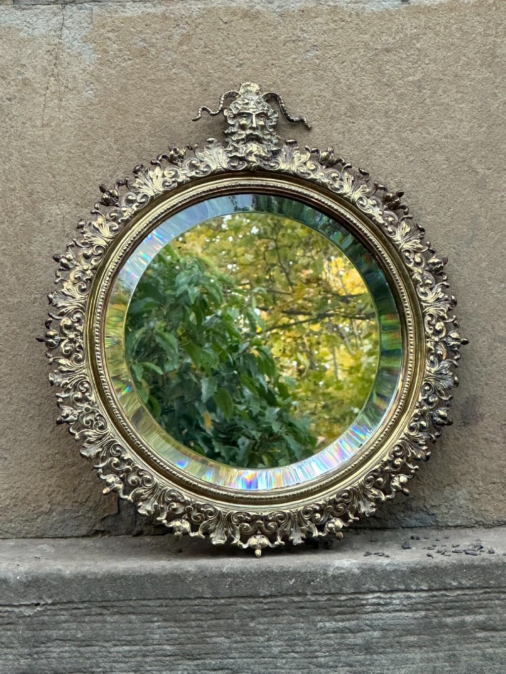 A circular Victorian cast brass wall mirror featuring a detailed crest with a mythological mask motif and a bevelled glass plate.
