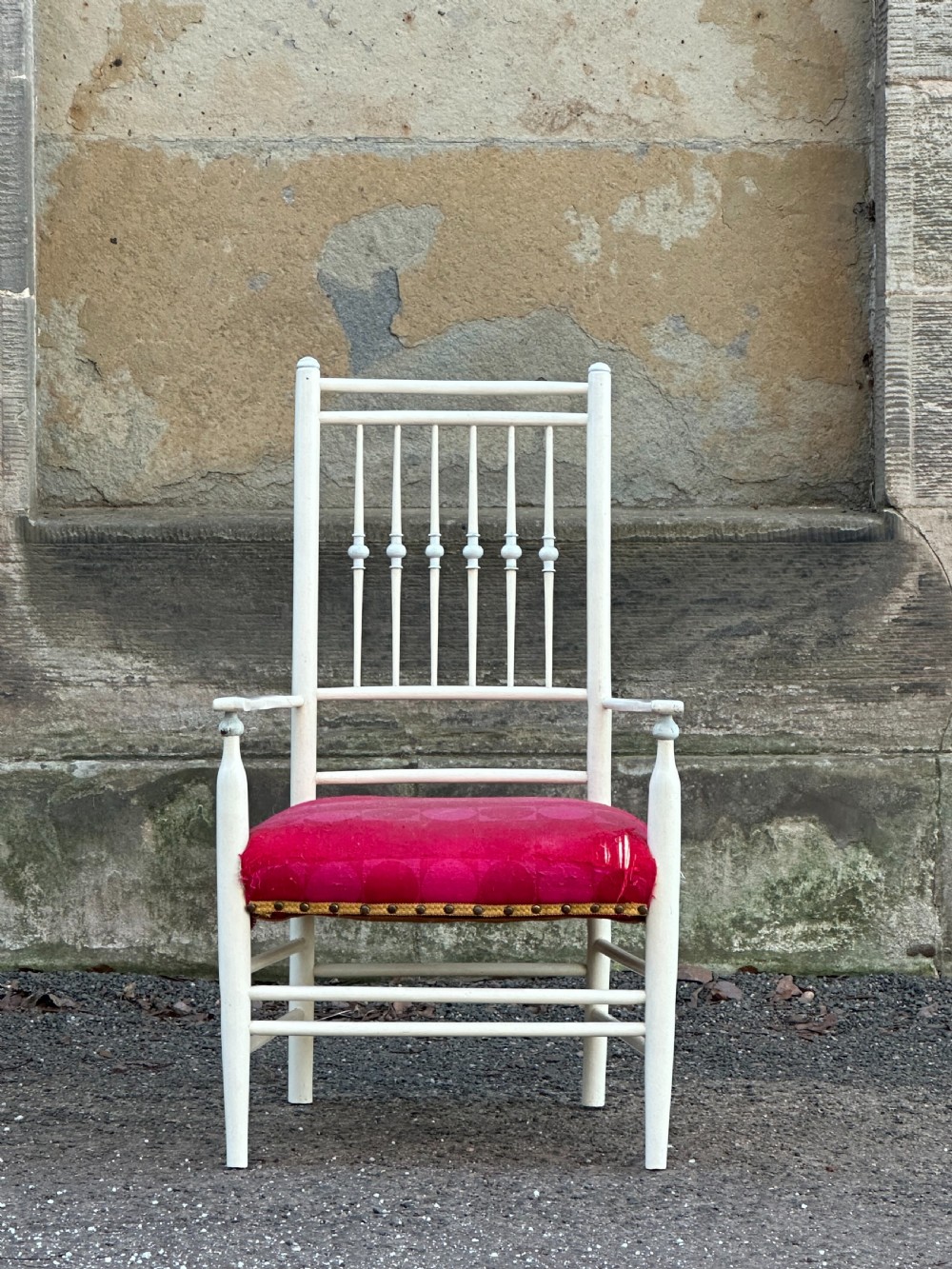 A white painted Arts and Crafts armchair with a tall spindle back, straight arms, and a padded red fabric seat secured with brass studs.