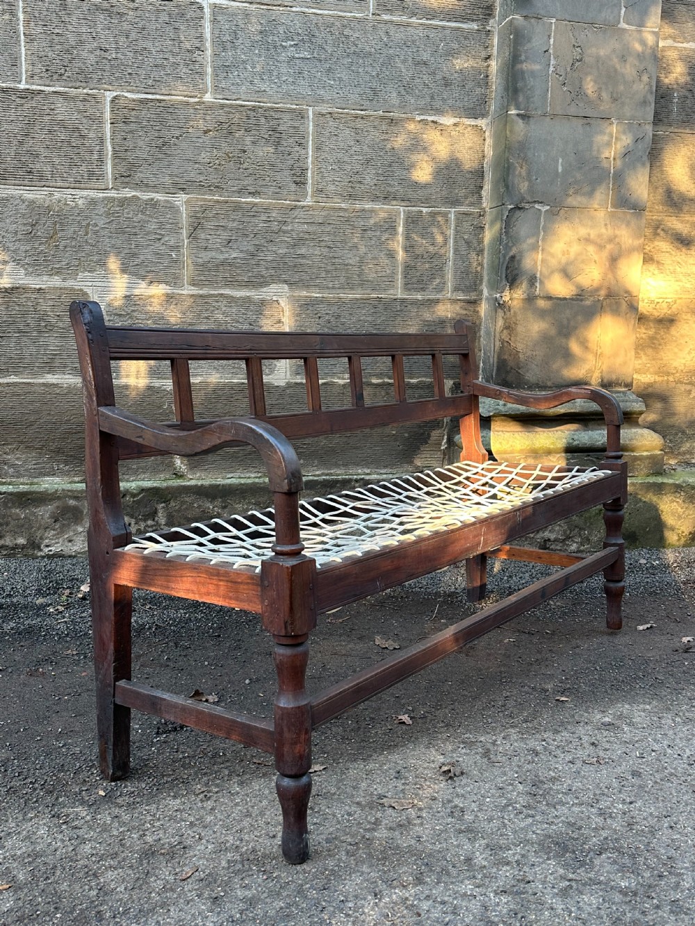 An early 19th-century South African stinkwood bench featuring a woven leather thong seat, curved armrests, and turned front legs. View X
