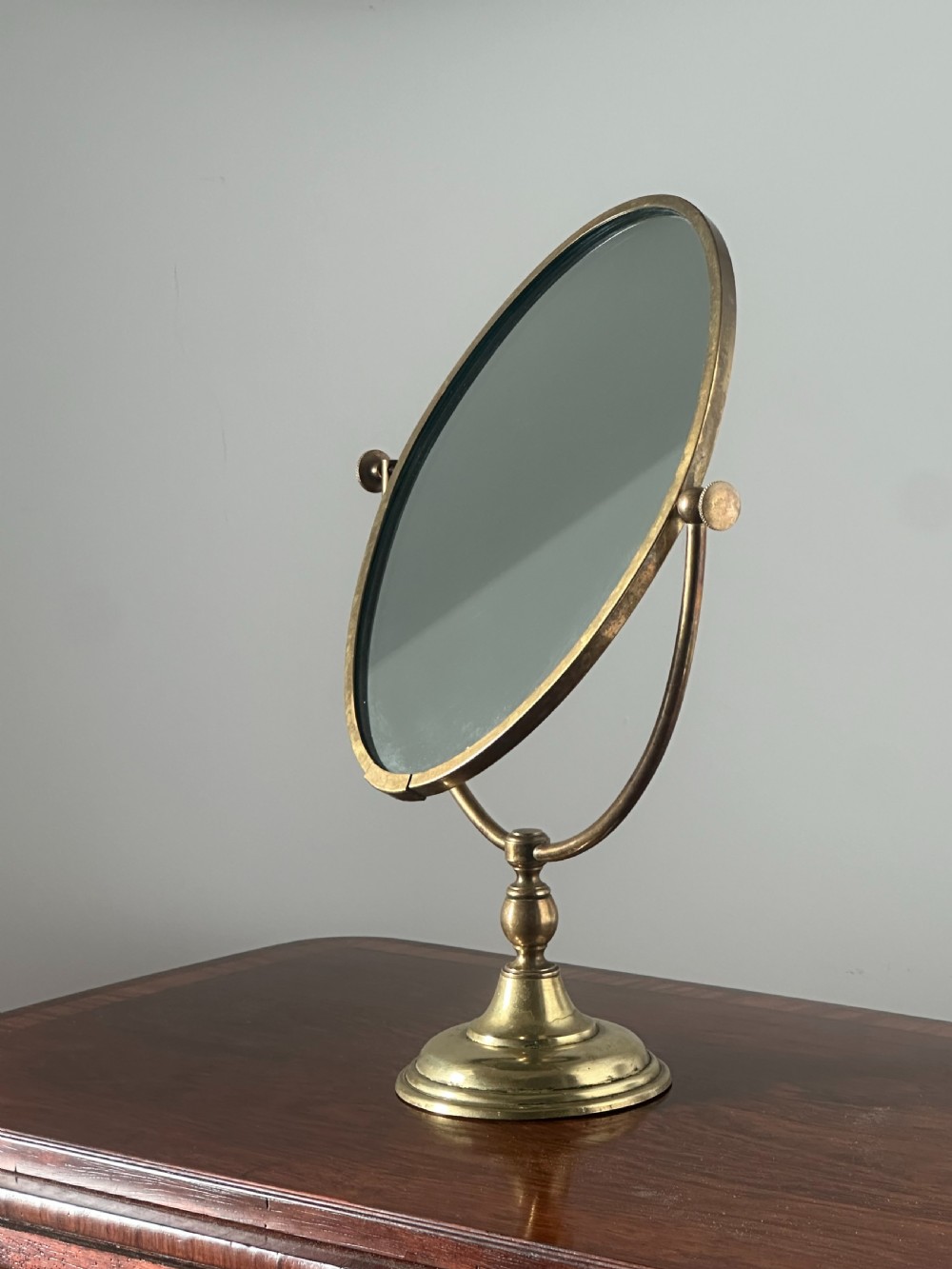 An oval brass shaving mirror on a matching stand sits atop a dark wooden surface. View IX