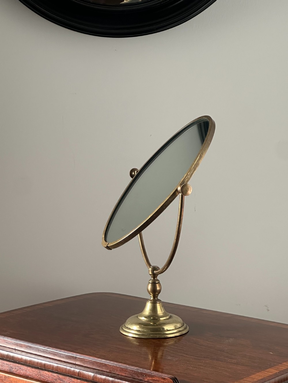 An oval brass shaving mirror on a matching stand sits atop a dark wooden surface.