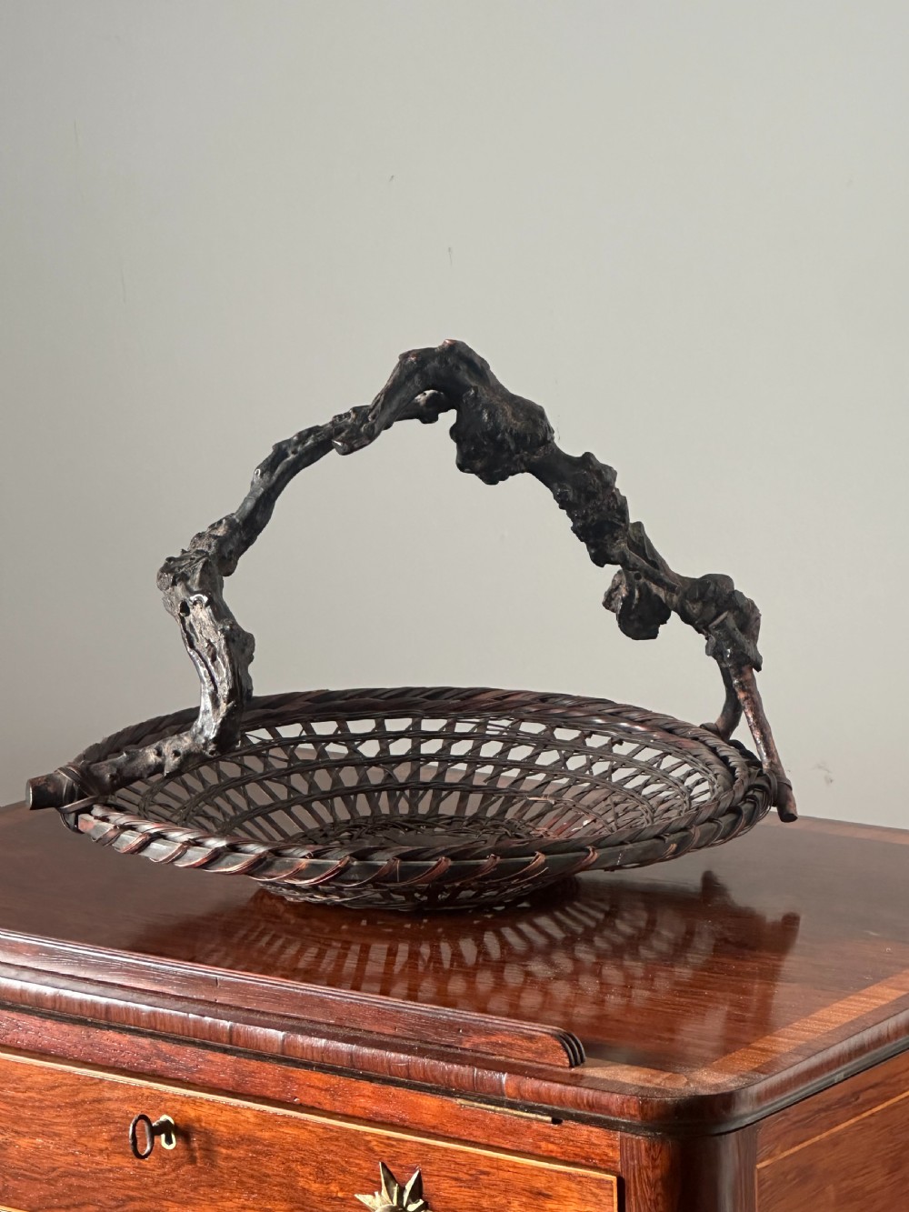 A shallow dark brown woven bamboo Ikebana flower basket featuring an organic gnarled natural rootwood overhead handle. View VII