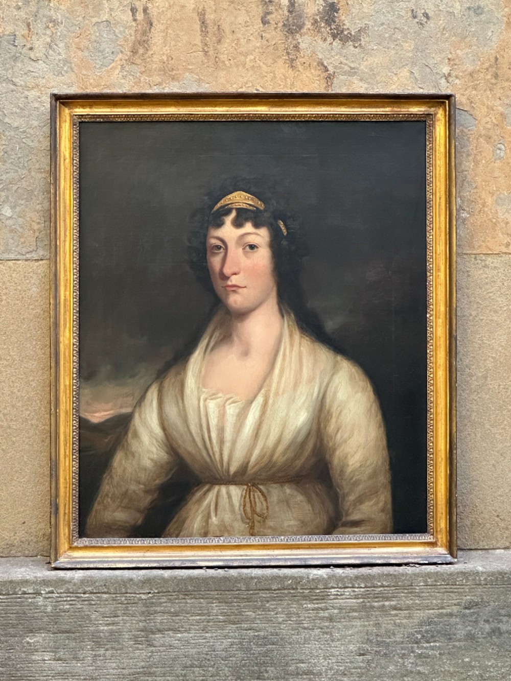 An early nineteenth-century portrait painting on canvas showing a woman in a cream Empire-line dress framed in carved gold leaf wood.