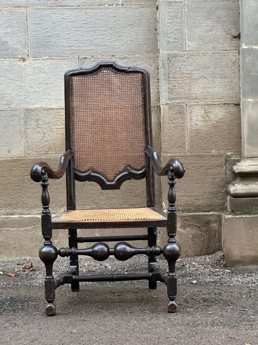 A dark painted walnut open armchair featuring a high woven cane back panel, scrolled arms, a cane seat, and bulbous turned stretchers.