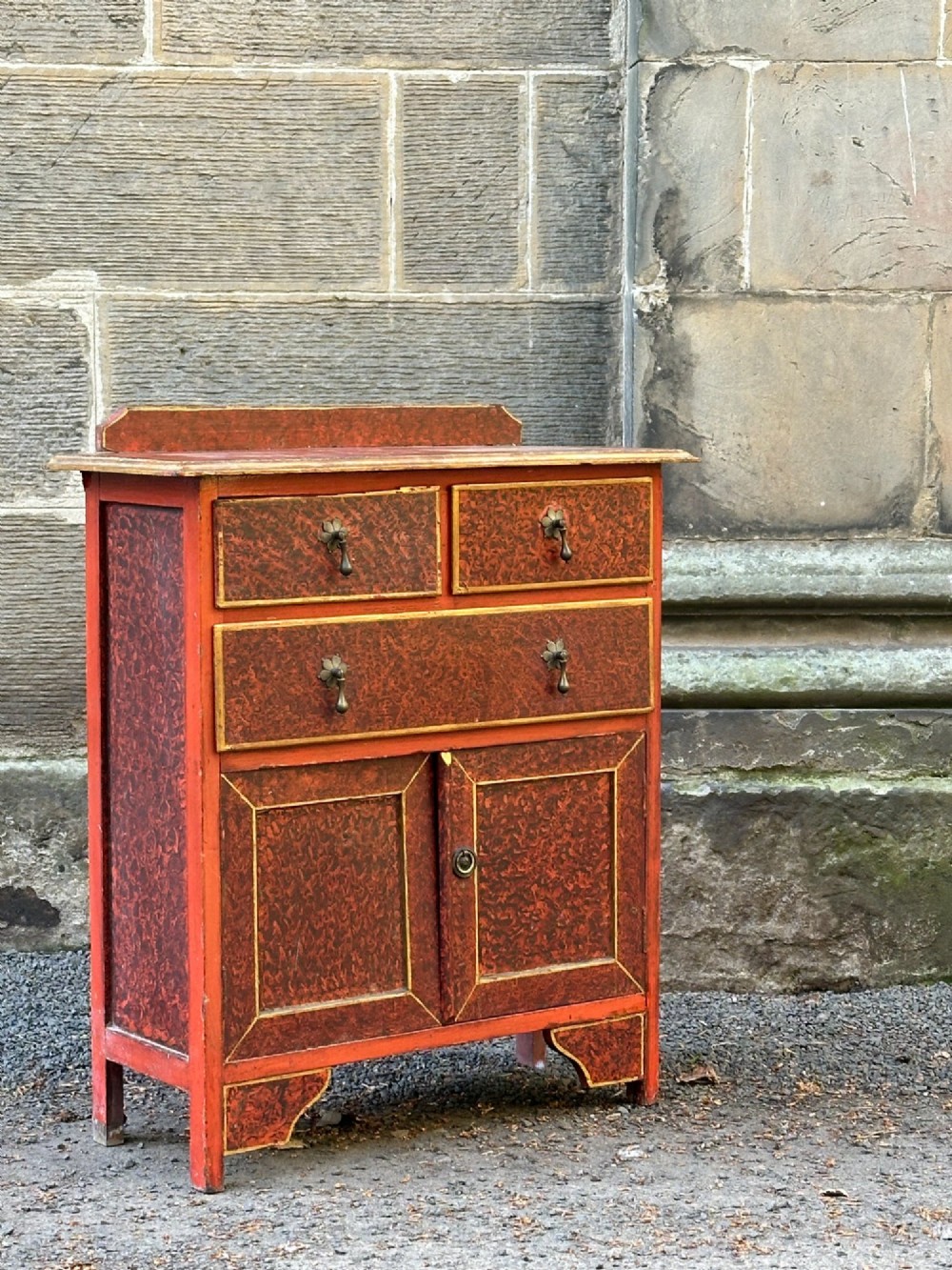 A red scumbled painted wooden side cabinet featuring three drawers above a two-door cupboard with gilded border trim and metal drop handles. View III