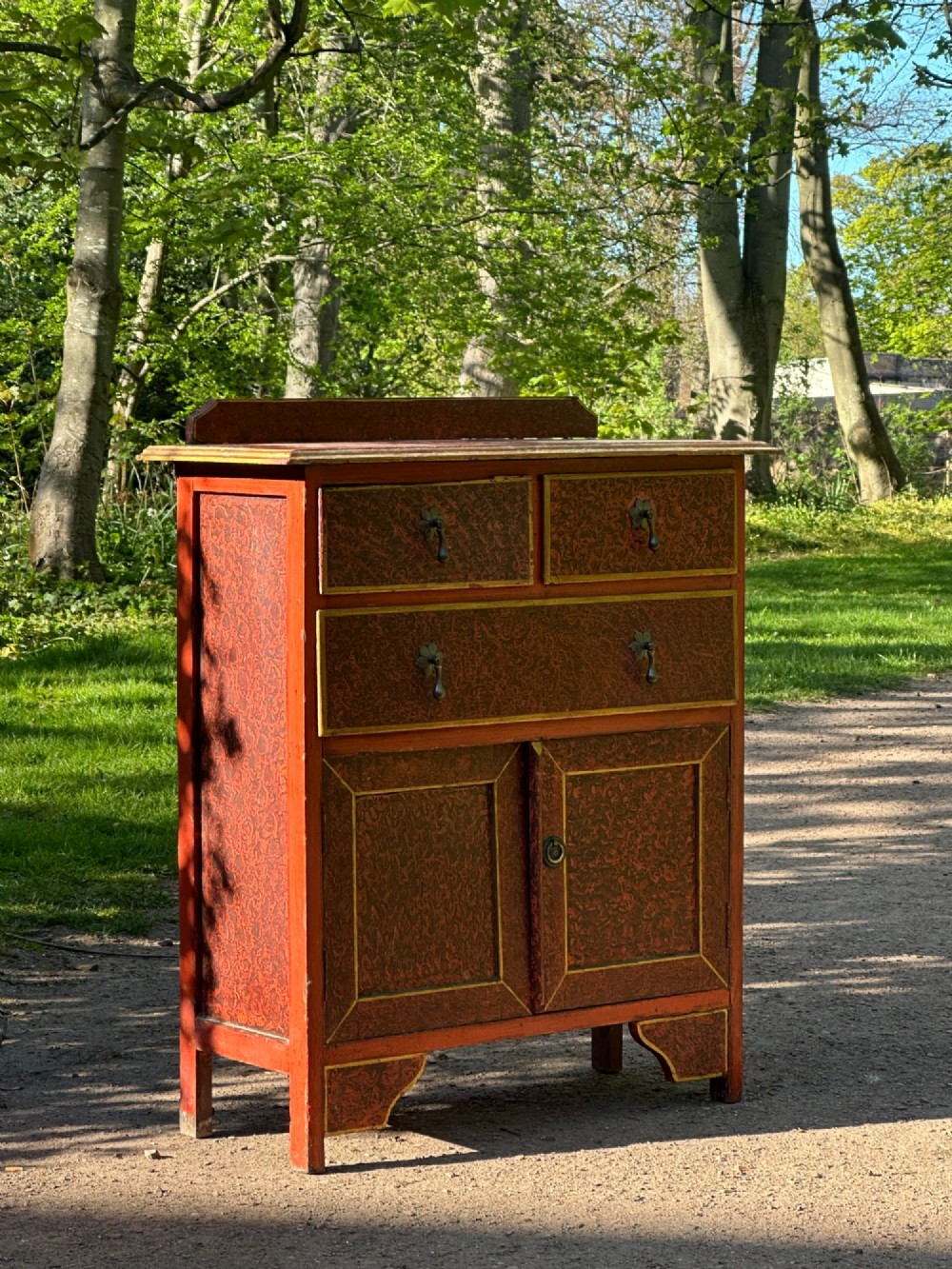 A red scumbled painted wooden side cabinet featuring three drawers above a two-door cupboard with gilded border trim and metal drop handles. View IV