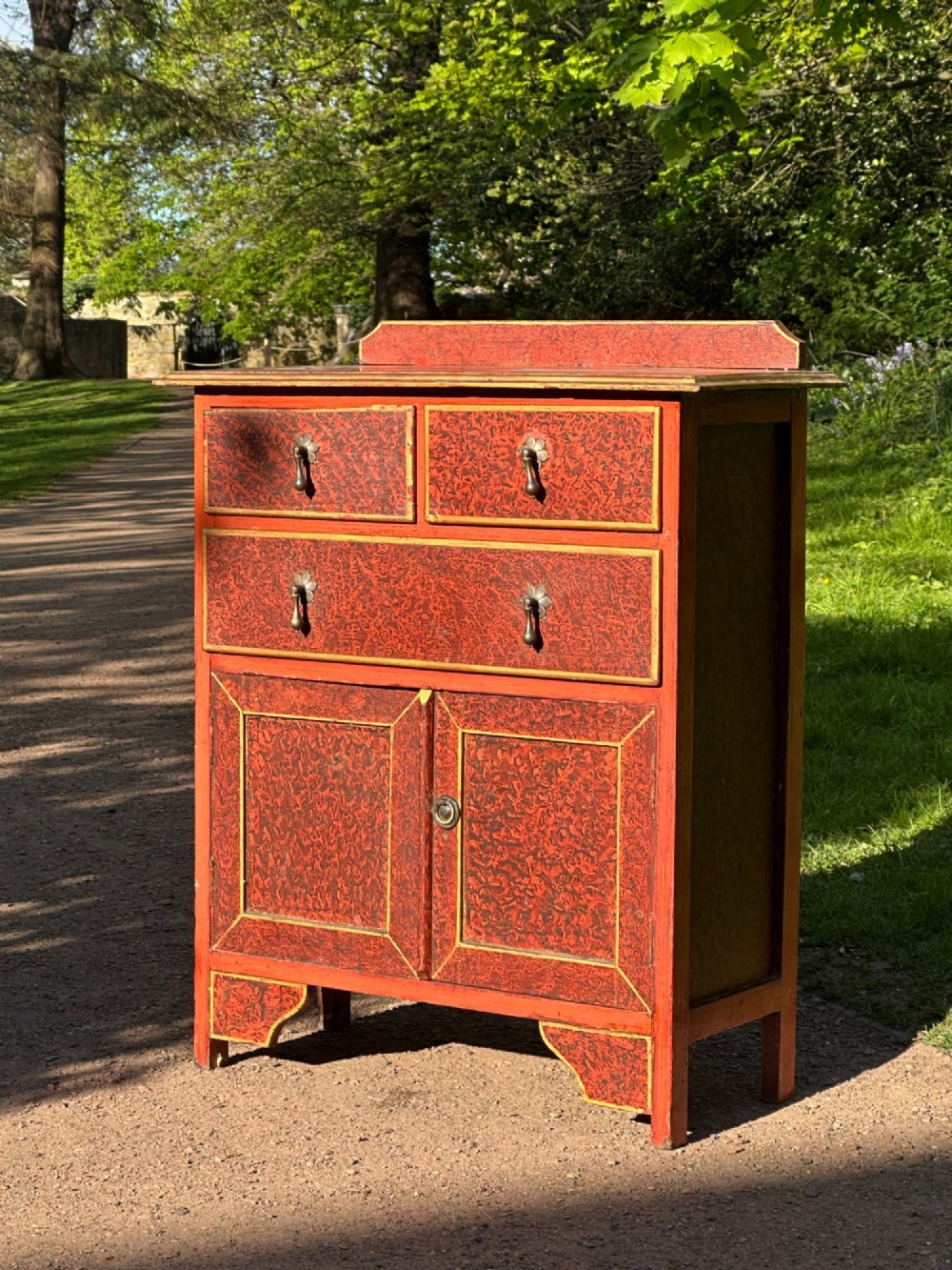 A red scumbled painted wooden side cabinet featuring three drawers above a two-door cupboard with gilded border trim and metal drop handles.