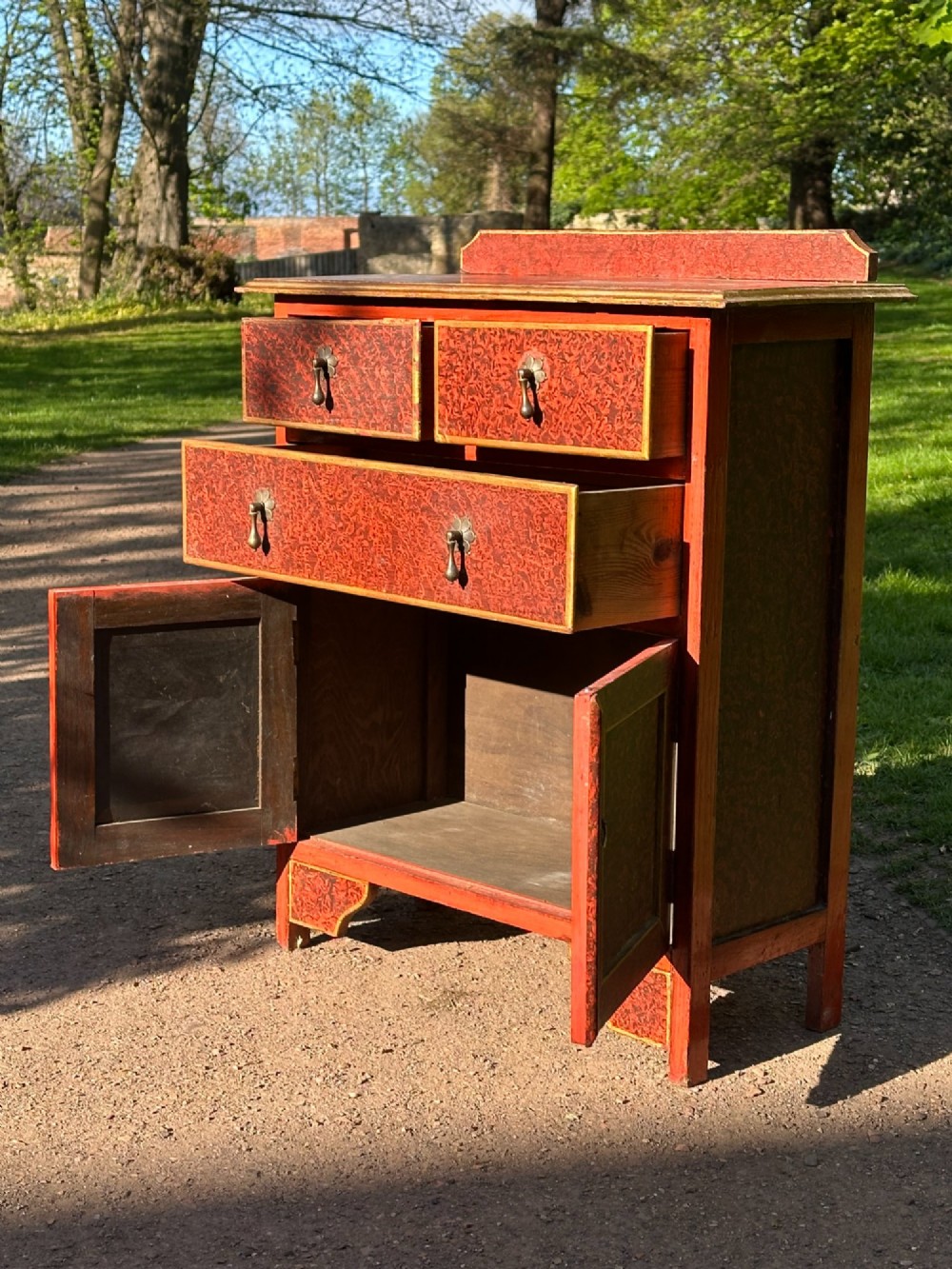 A red scumbled painted wooden side cabinet featuring three drawers above a two-door cupboard with gilded border trim and metal drop handles. View VIII