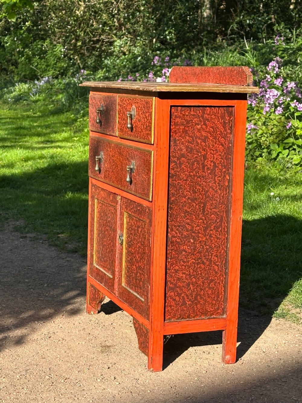 A red scumbled painted wooden side cabinet featuring three drawers above a two-door cupboard with gilded border trim and metal drop handles. View IX