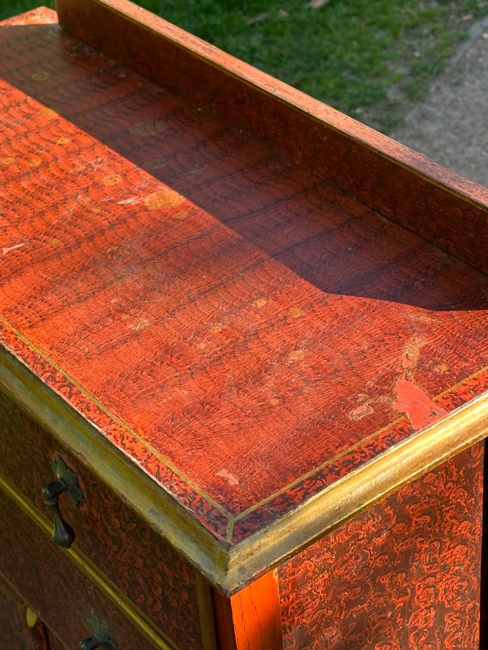 A red scumbled painted wooden side cabinet featuring three drawers above a two-door cupboard with gilded border trim and metal drop handles. View X