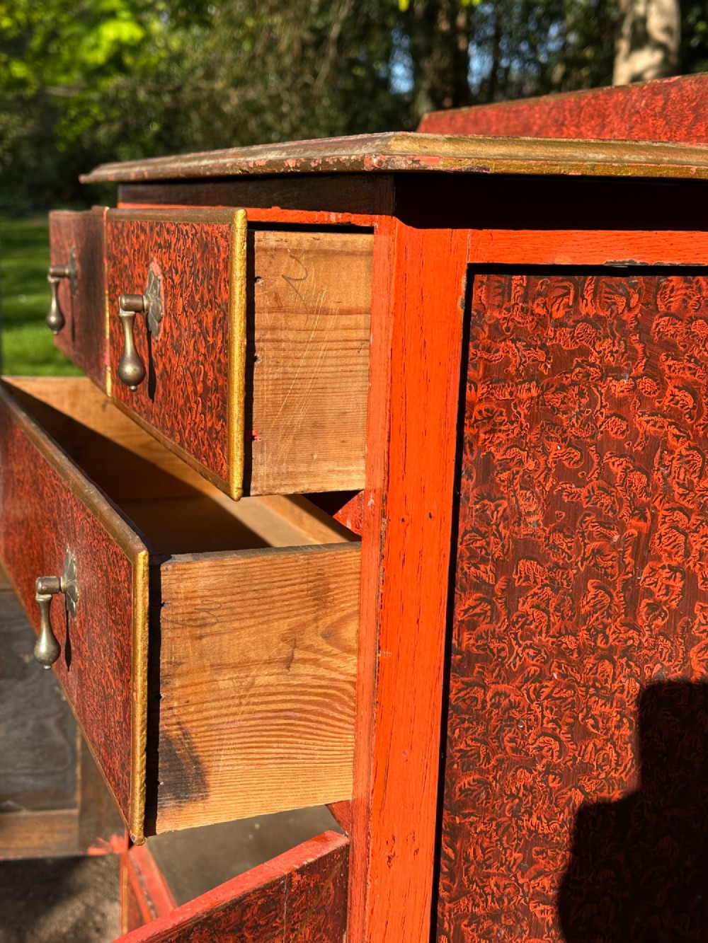 A red scumbled painted wooden side cabinet featuring three drawers above a two-door cupboard with gilded border trim and metal drop handles. View XII