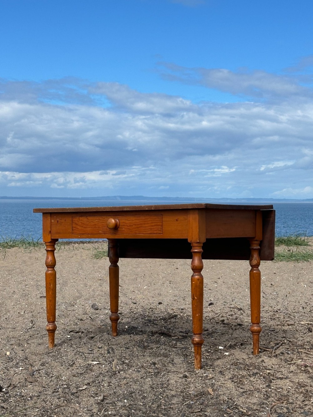 A 19th-century pine kitchen preparation table featuring turned legs, a single frieze drawer with a wooden knob, and a drop-leaf extension.