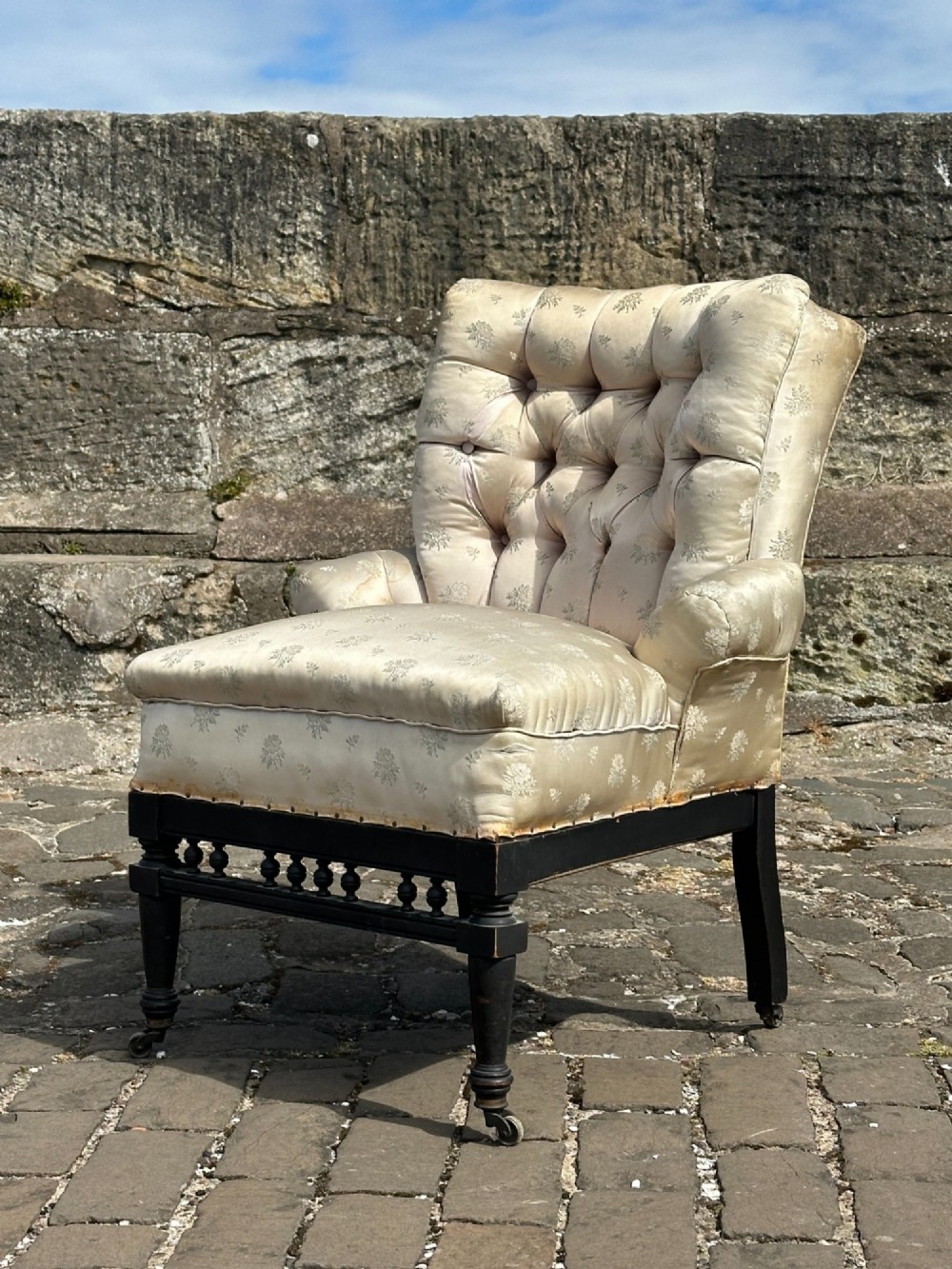 A Victorian button-back drawing room chair with cream damask upholstery, ebonised turned legs, a spindled stretcher gallery, and brass casters.