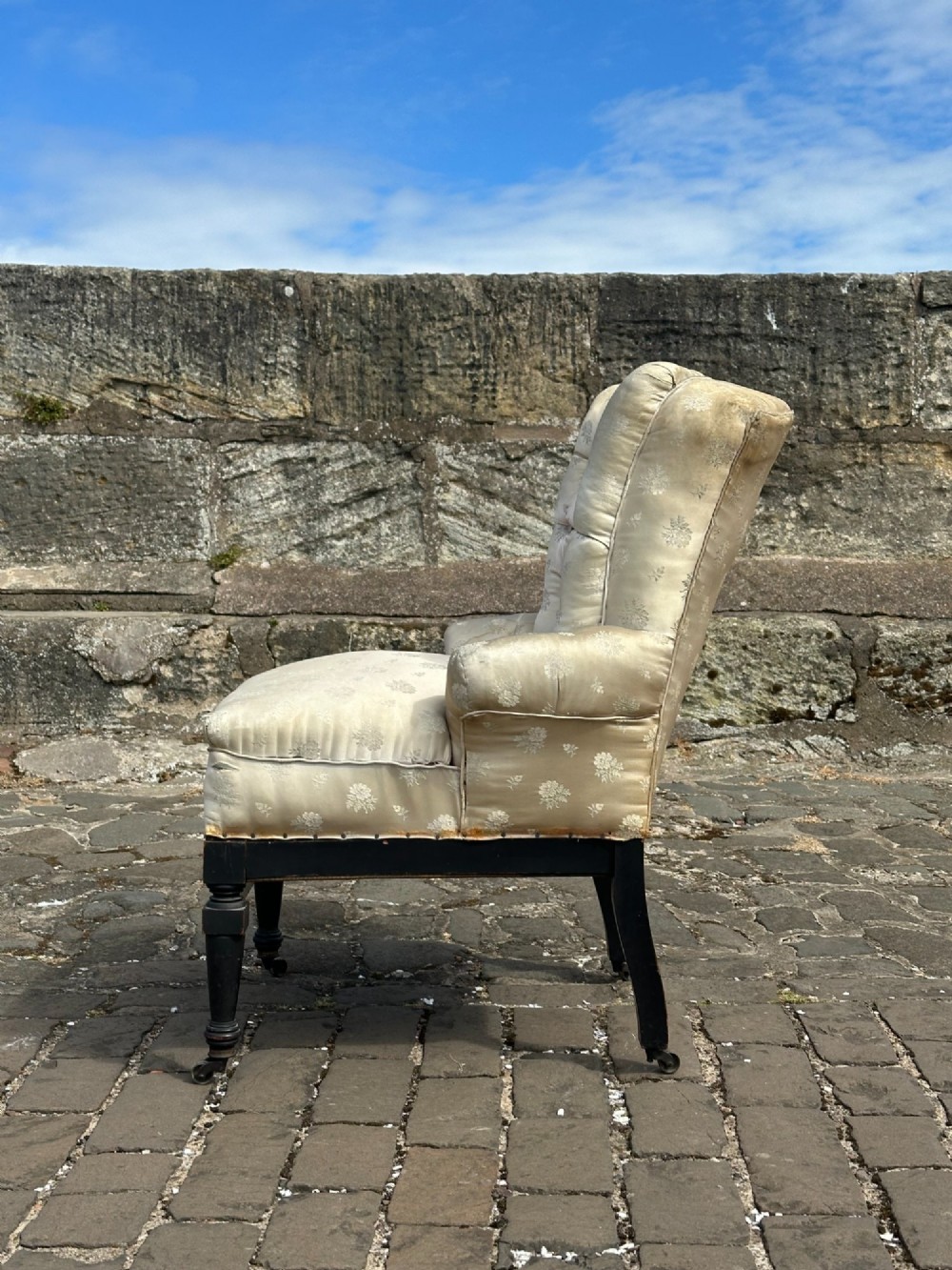 A Victorian button-back drawing room chair with cream damask upholstery, ebonised turned legs, a spindled stretcher gallery, and brass casters. View V