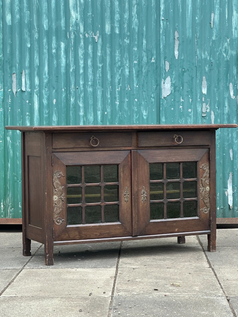 A dark oak Arts and Crafts side cabinet with two upper drawers, lattice glazed double doors, and decorative brass mounts.