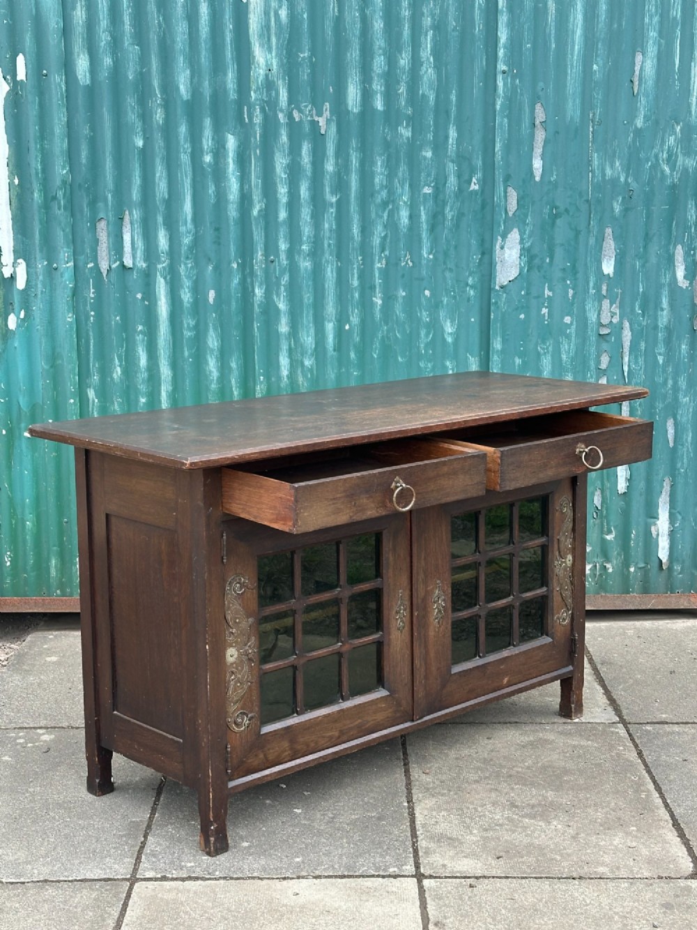 A dark oak Arts and Crafts side cabinet with two upper drawers, lattice glazed double doors, and decorative brass mounts. View II