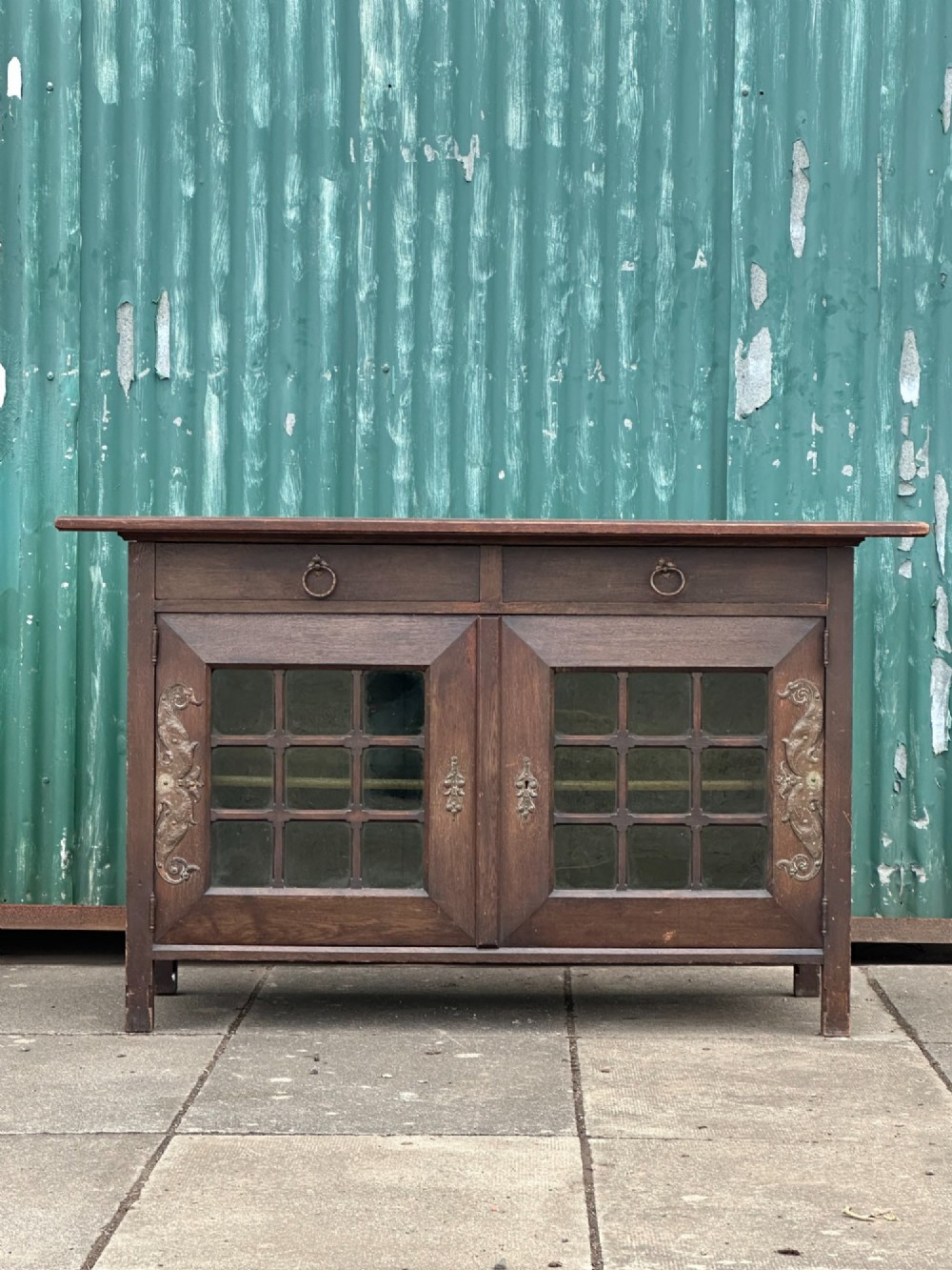 A dark oak Arts and Crafts side cabinet with two upper drawers, lattice glazed double doors, and decorative brass mounts. View III