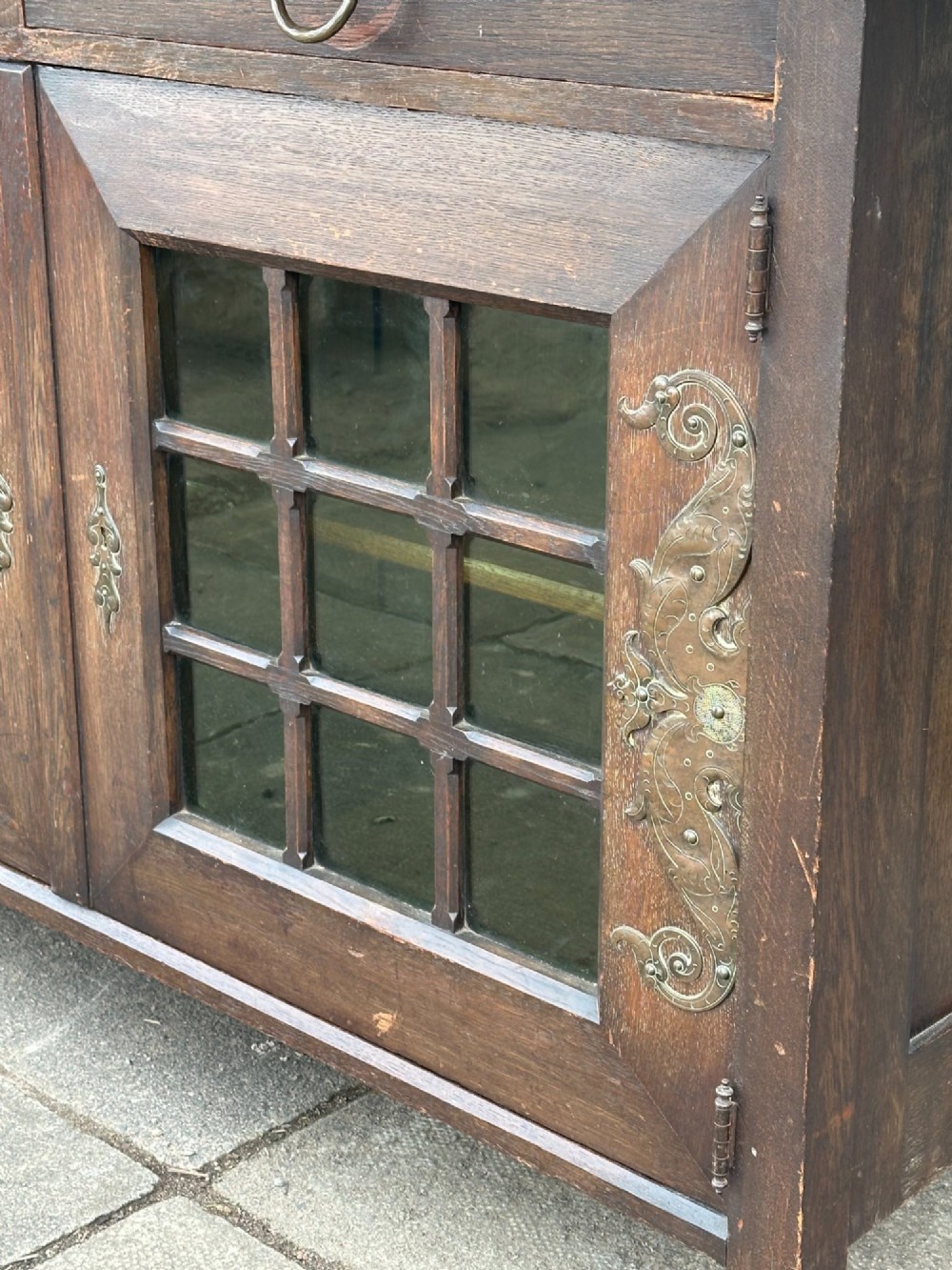 A dark oak Arts and Crafts side cabinet with two upper drawers, lattice glazed double doors, and decorative brass mounts. View IV