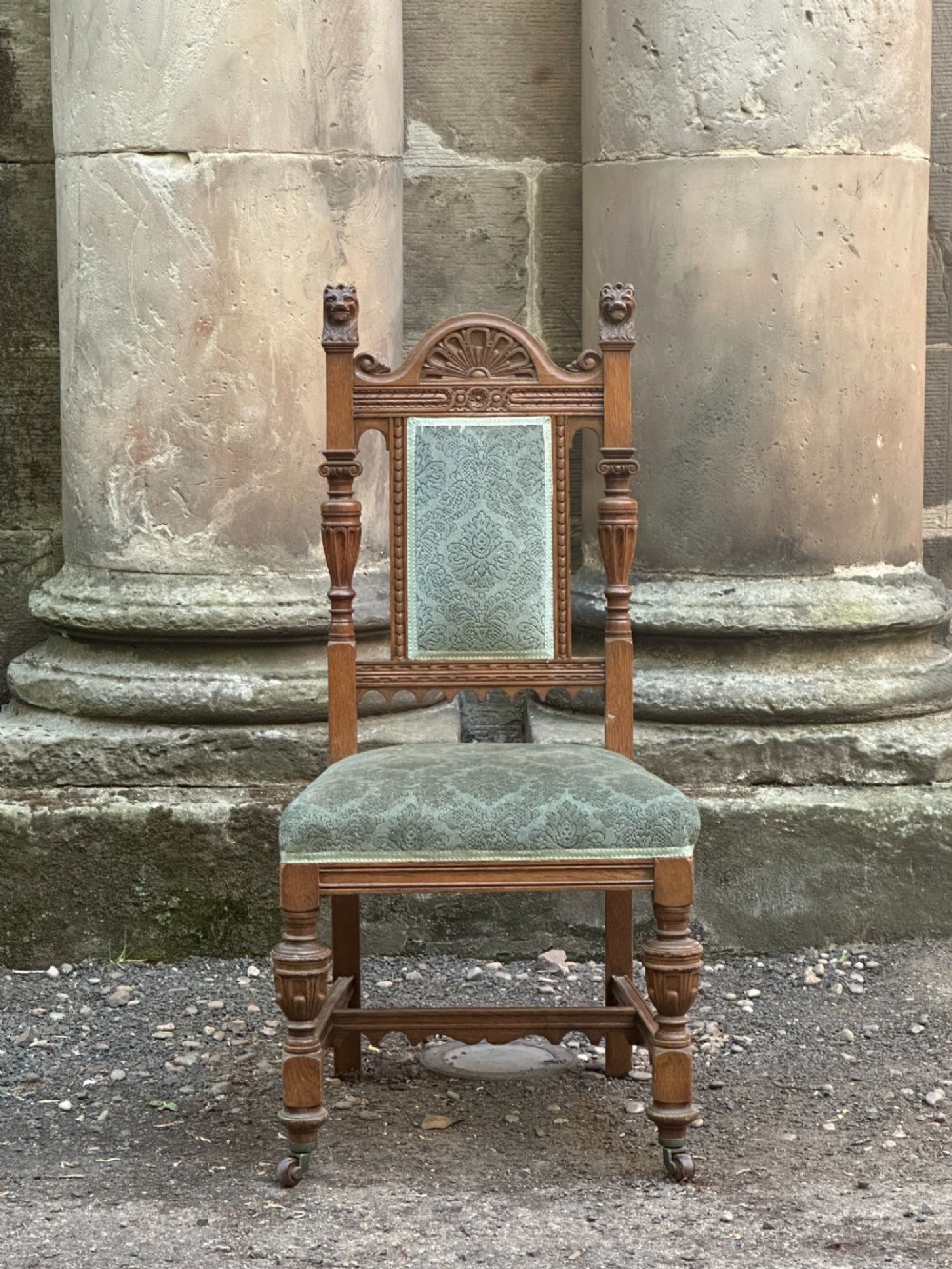 A carved oak hall chair featuring lion mask finials, shell cresting, turned front legs on castors, and sage green pattern upholstery. View II