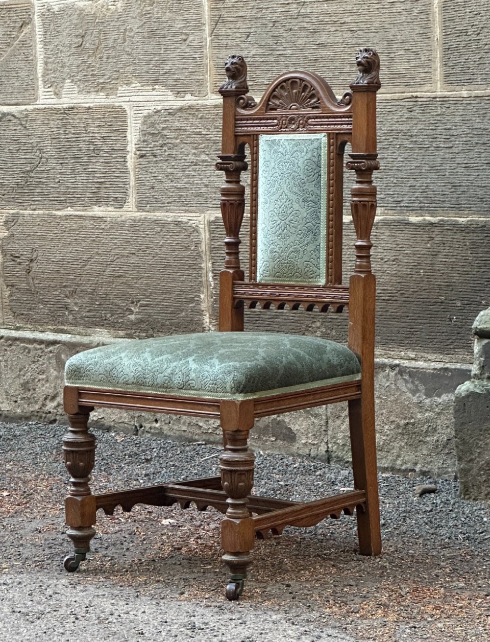 A carved oak hall chair featuring lion mask finials, shell cresting, turned front legs on castors, and sage green pattern upholstery.