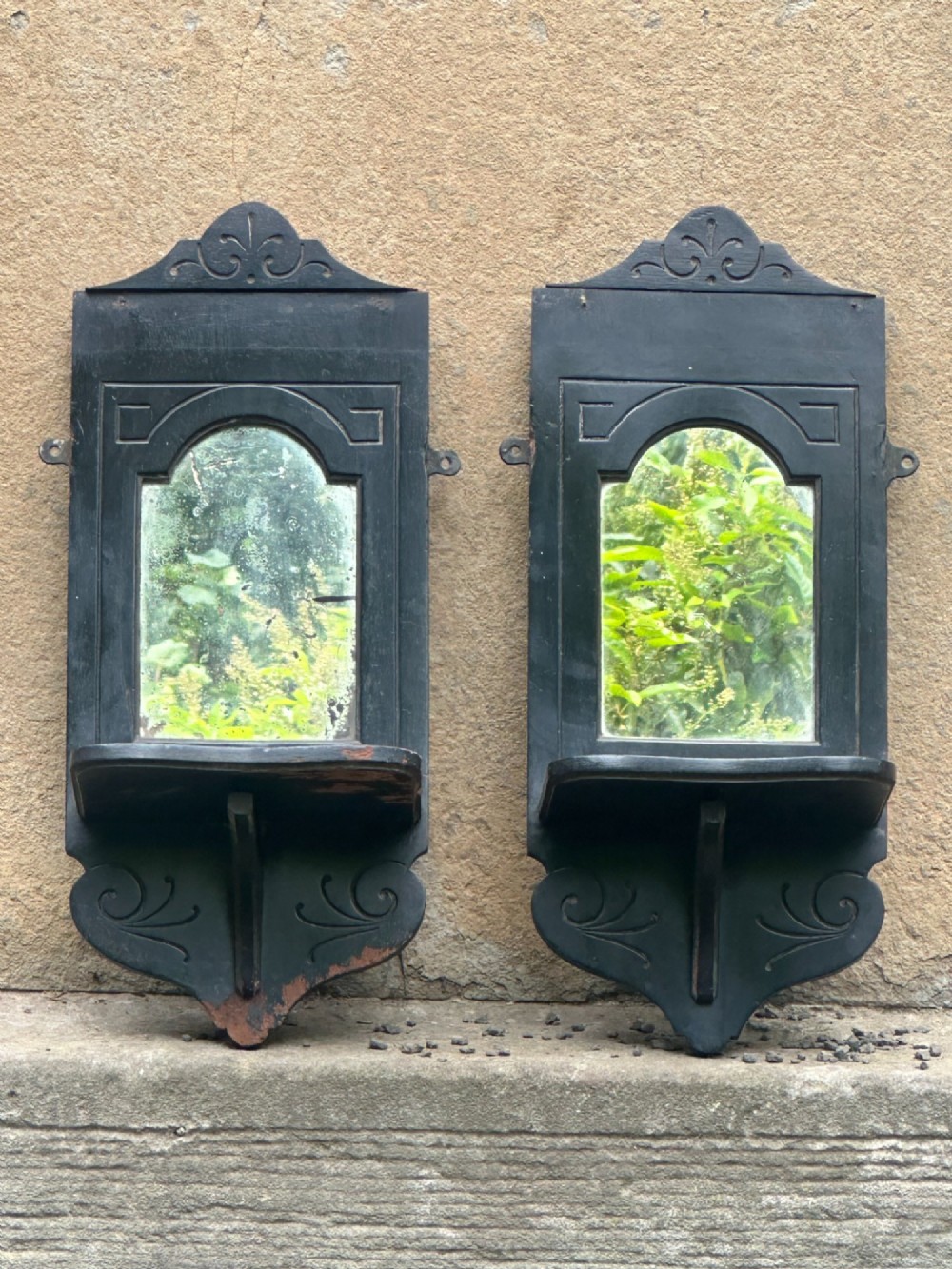 A pair of late 19th-century black ebonised wooden wall girandoles with arched mirror plates, incised scroll detailing, and integrated shelves.