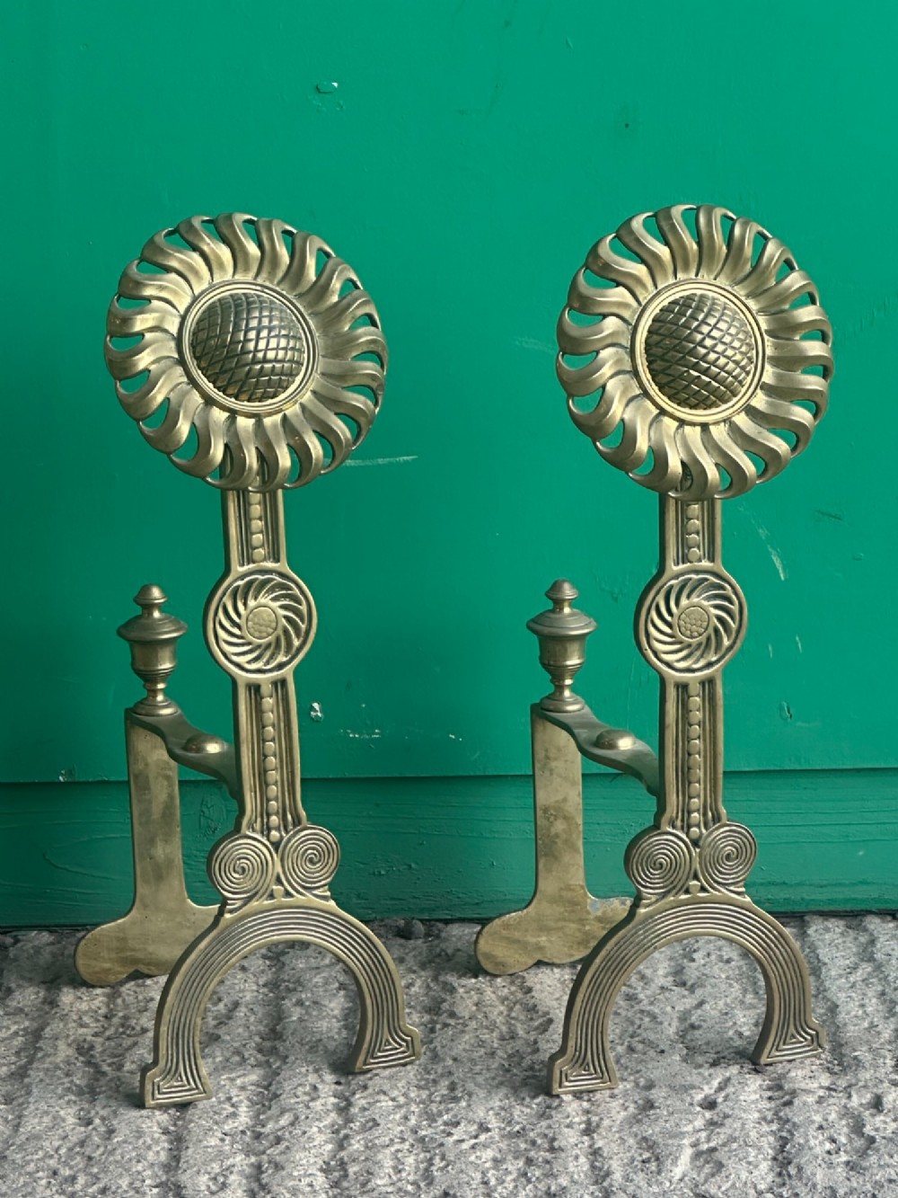 A pair of late Victorian cast brass andirons featuring stylised sunflower tops and geometric scroll detailing.