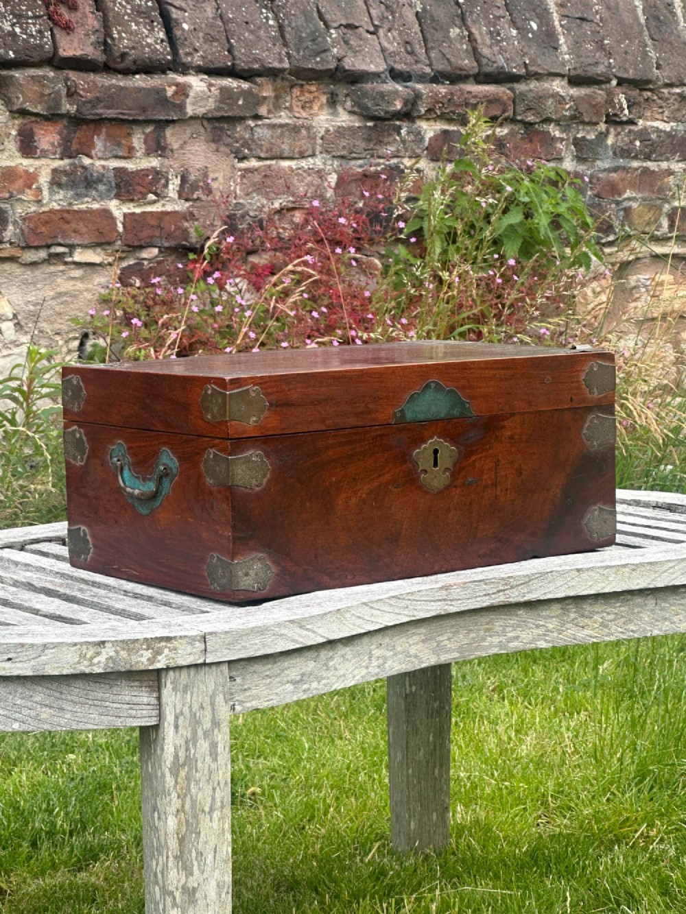 A rectangular reddish-brown padouk wood deed box featuring brass corner mounts, a central keyhole escutcheon, and brass side carrying handles.