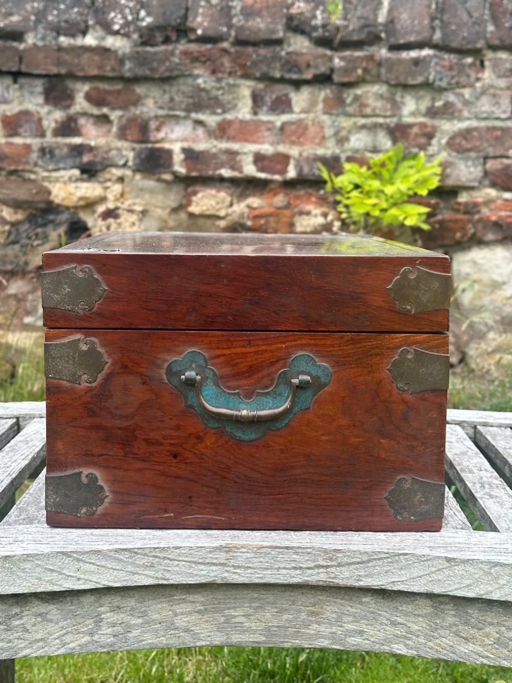 A rectangular reddish-brown padouk wood deed box featuring brass corner mounts, a central keyhole escutcheon, and brass side carrying handles. View V