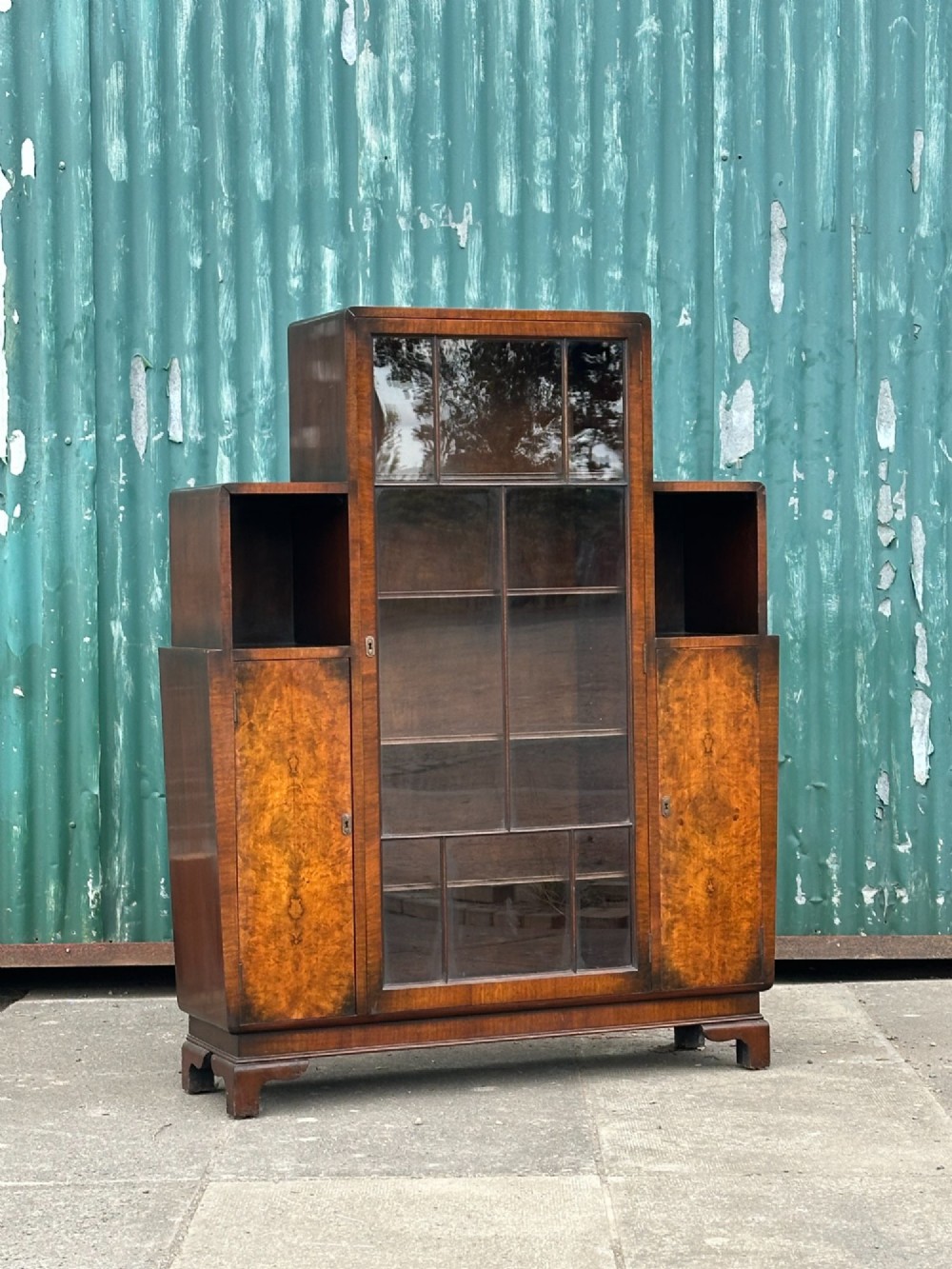 An architectural 1930s Art Deco cabinet made of warm brown walnut wood with a tall central glazed door flanked by lower cupboard doors and open alcoves.