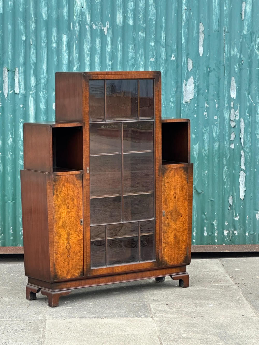 An architectural 1930s Art Deco cabinet made of warm brown walnut wood with a tall central glazed door flanked by lower cupboard doors and open alcoves. View II