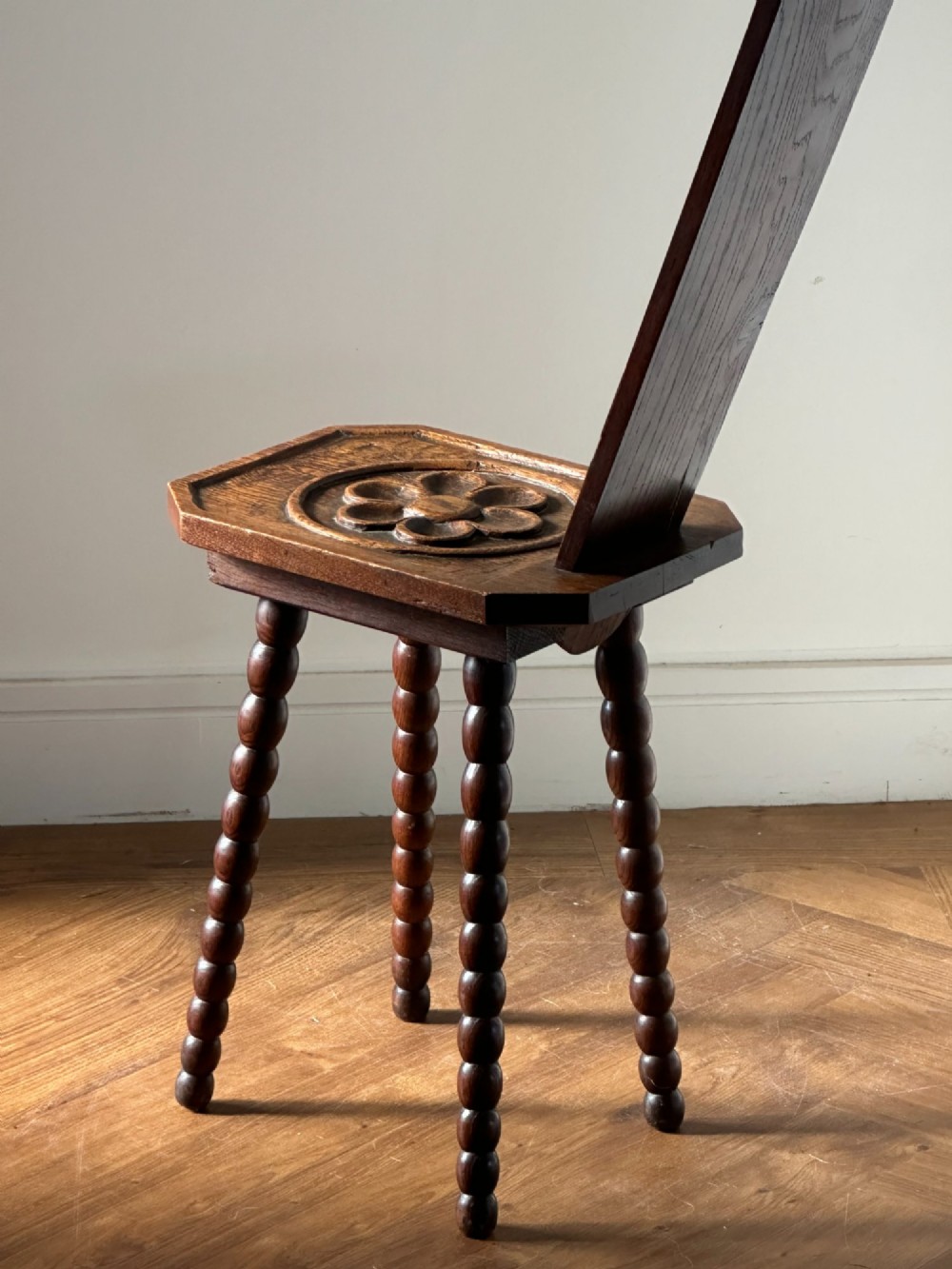 An Arts and Crafts carved wooden spinning stool featuring a tall narrow backrest, a relief-carved floral seat, and bobbin-turned legs. View II