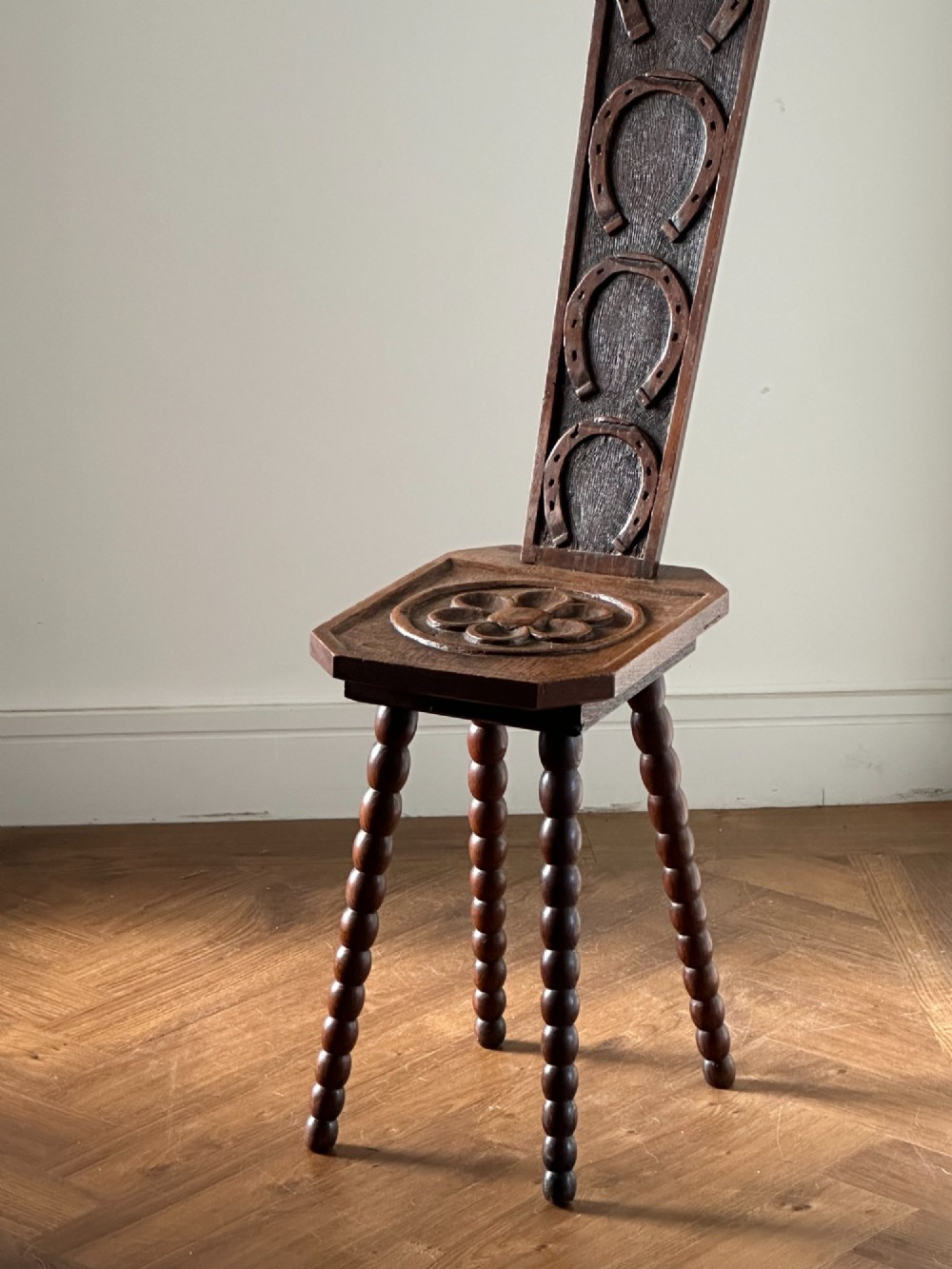 An Arts and Crafts carved wooden spinning stool featuring a tall narrow backrest, a relief-carved floral seat, and bobbin-turned legs. View V