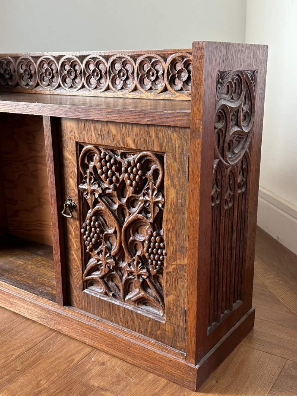 A low Arts and Crafts carved oak table cabinet featuring an open left compartment, a carved door on the right with foliate motifs, and a gallery top with carved rosettes. View V