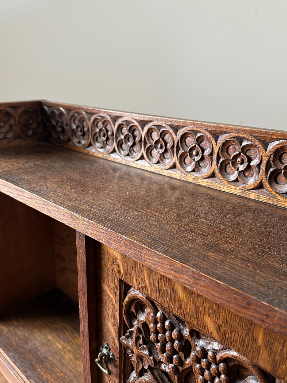 A low Arts and Crafts carved oak table cabinet featuring an open left compartment, a carved door on the right with foliate motifs, and a gallery top with carved rosettes. View VI