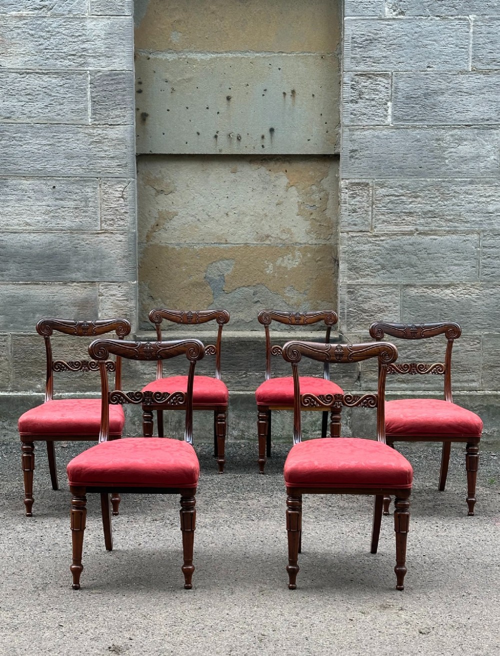 Set of six antique George IV rosewood dining chairs featuring carved top rails, horizontal back splats, turned front legs, and red upholstered seats.