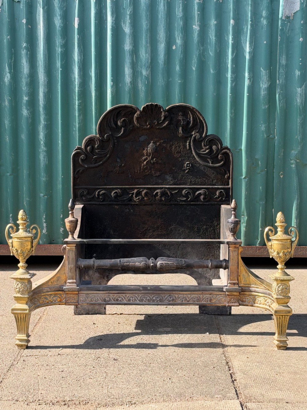 A late nineteenth-century George III style fire grate featuring an ornate cast iron fireback with scrollwork and decorative brass urn finials set on tapered brass legs.