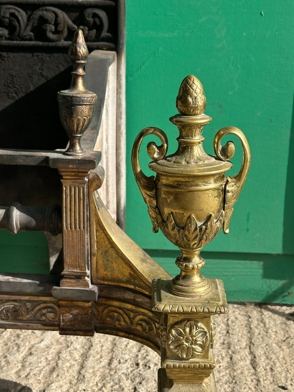 A late nineteenth-century George III style fire grate featuring an ornate cast iron fireback with scrollwork and decorative brass urn finials set on tapered brass legs. View III