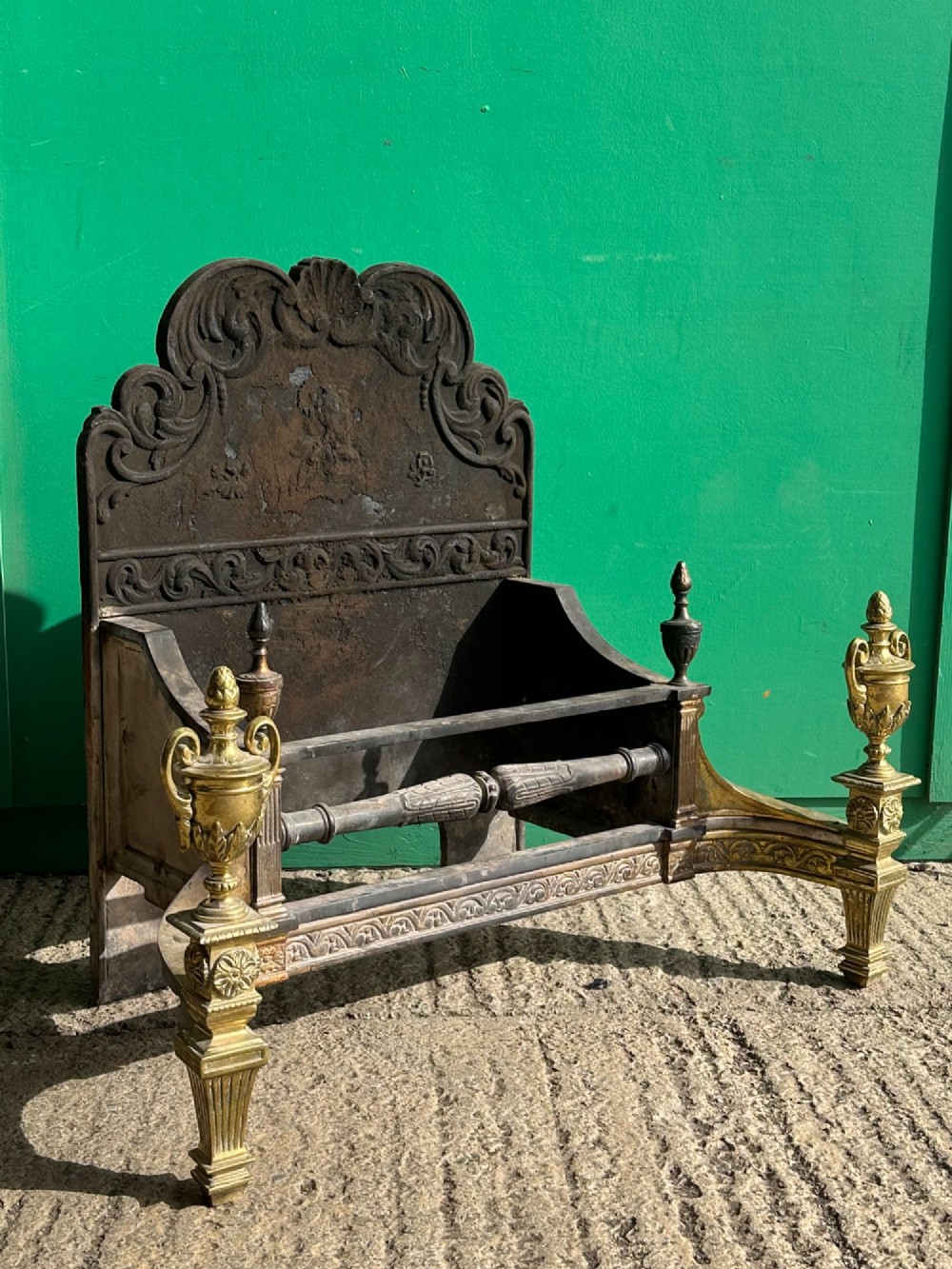 A late nineteenth-century George III style fire grate featuring an ornate cast iron fireback with scrollwork and decorative brass urn finials set on tapered brass legs. View IX