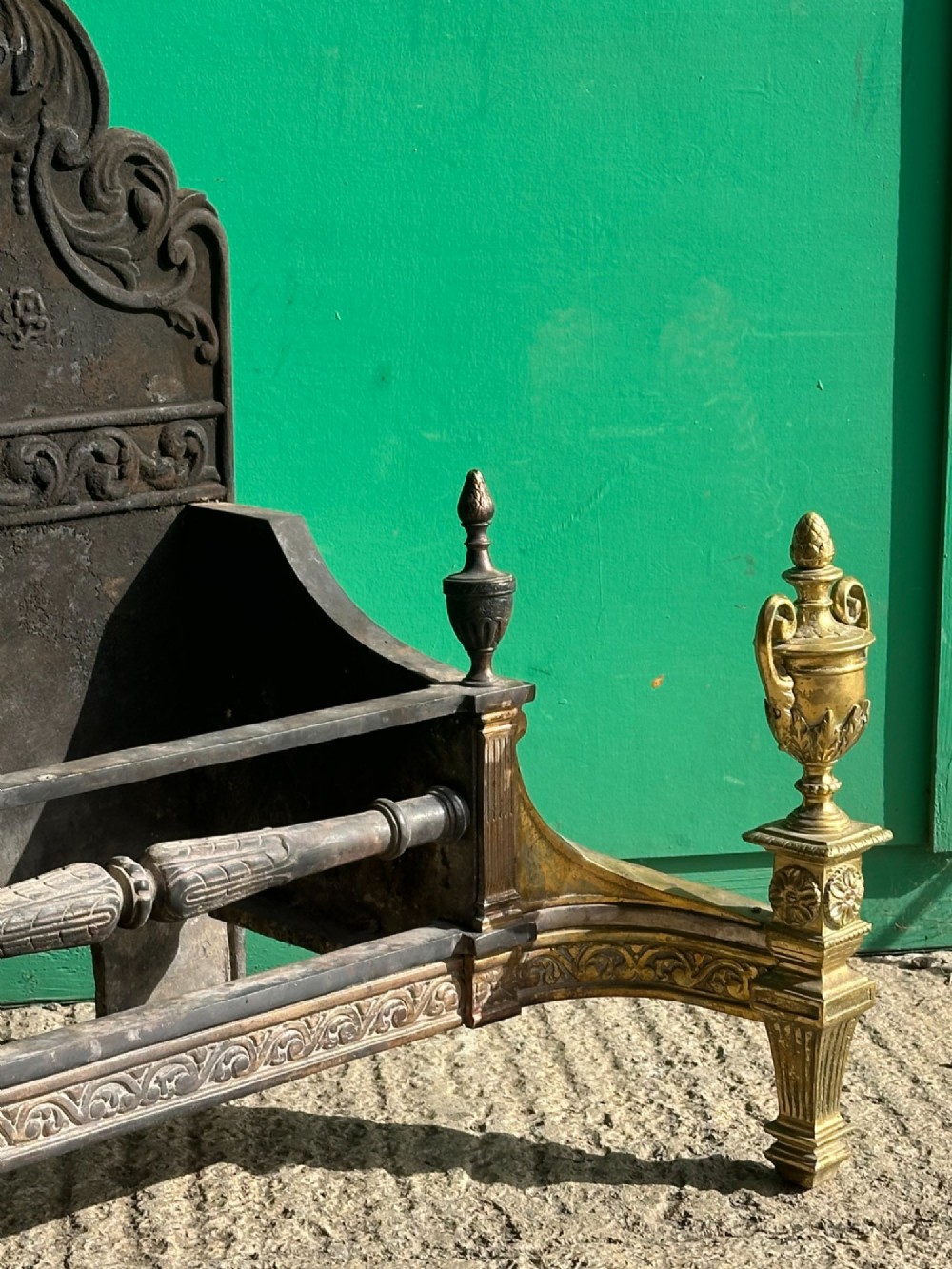 A late nineteenth-century George III style fire grate featuring an ornate cast iron fireback with scrollwork and decorative brass urn finials set on tapered brass legs. View X